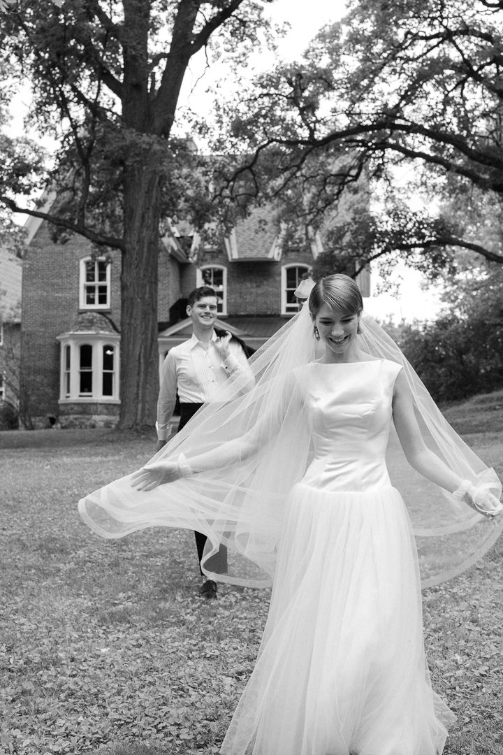 Black and white photo of a smiling bride in a flowing veil and gown with groom walking behind her outdoors.