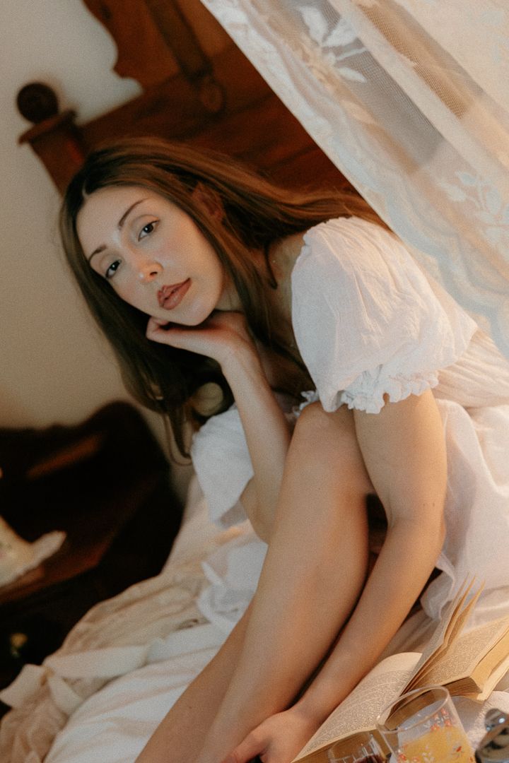 Young woman in a white puff-sleeve dress sitting on a bed surrounded by soft white linens and an open book.