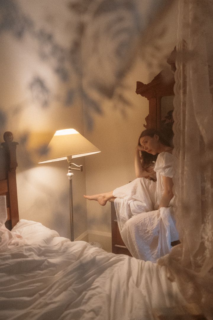 Woman in white dress sitting on bed beside glowing lamp in vintage bedroom with floral wallpaper.