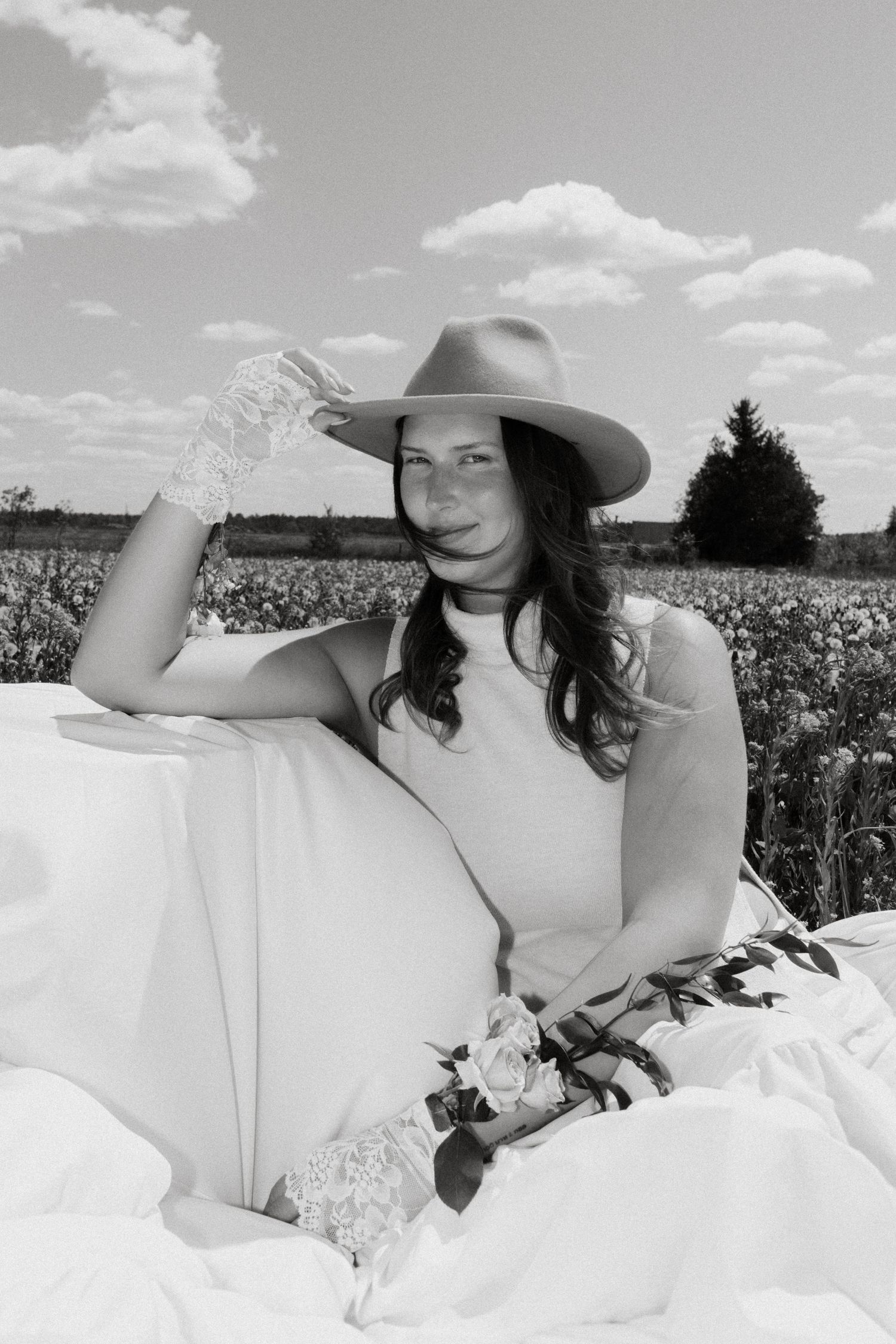 Black and white photo of a bride in a white dress and cowboy hat holding roses in a blooming field.