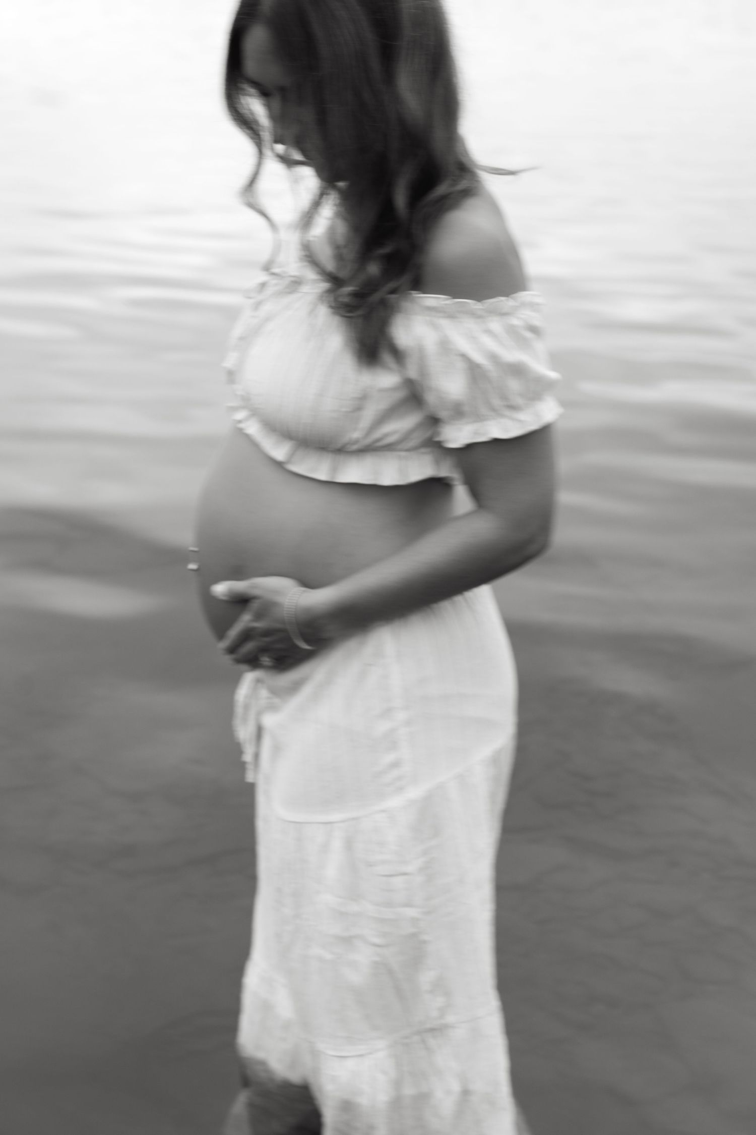 Black and white maternity photo of pregnant woman in white off-shoulder dress standing in water, cradling her baby bump.