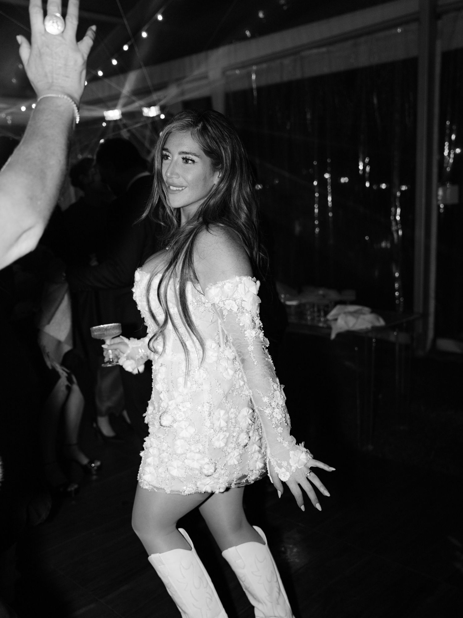 Black and white photo of bride in short floral dress and white boots dancing at wedding reception.