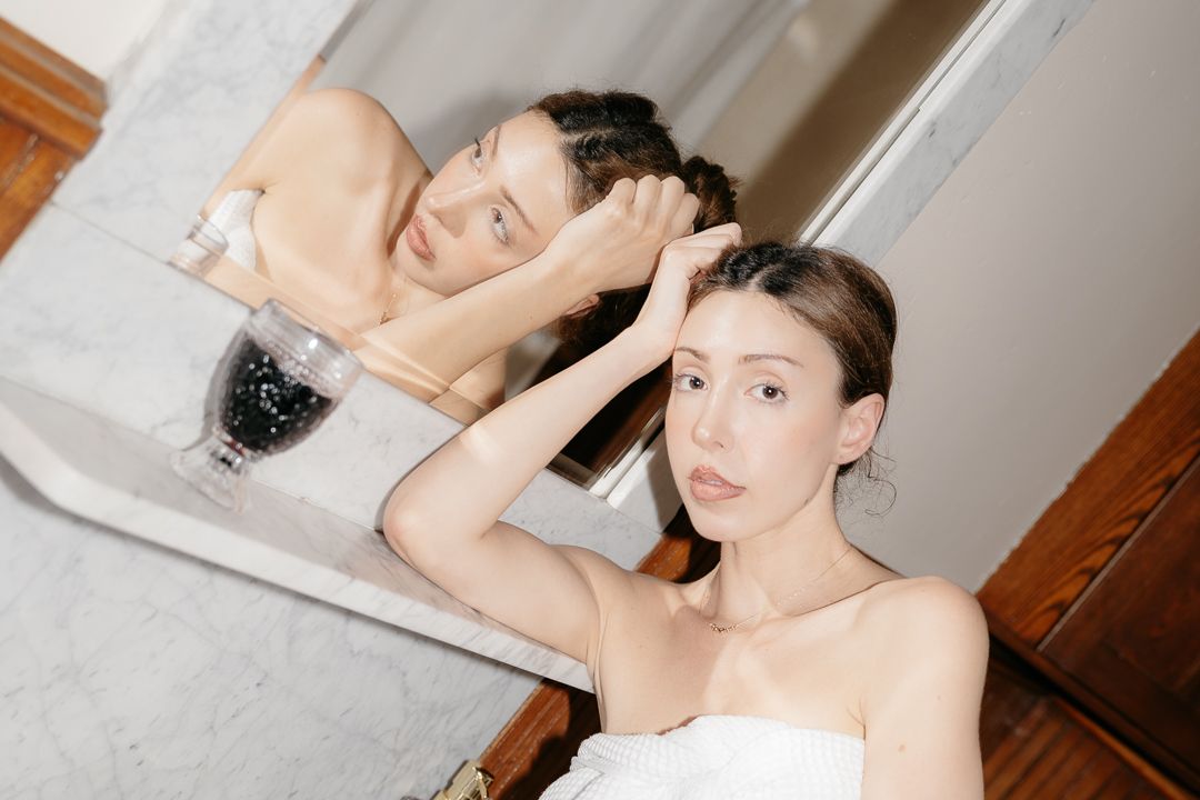 Woman adjusting her hair while looking in a bathroom mirror, with a wine glass visible on the counter.
