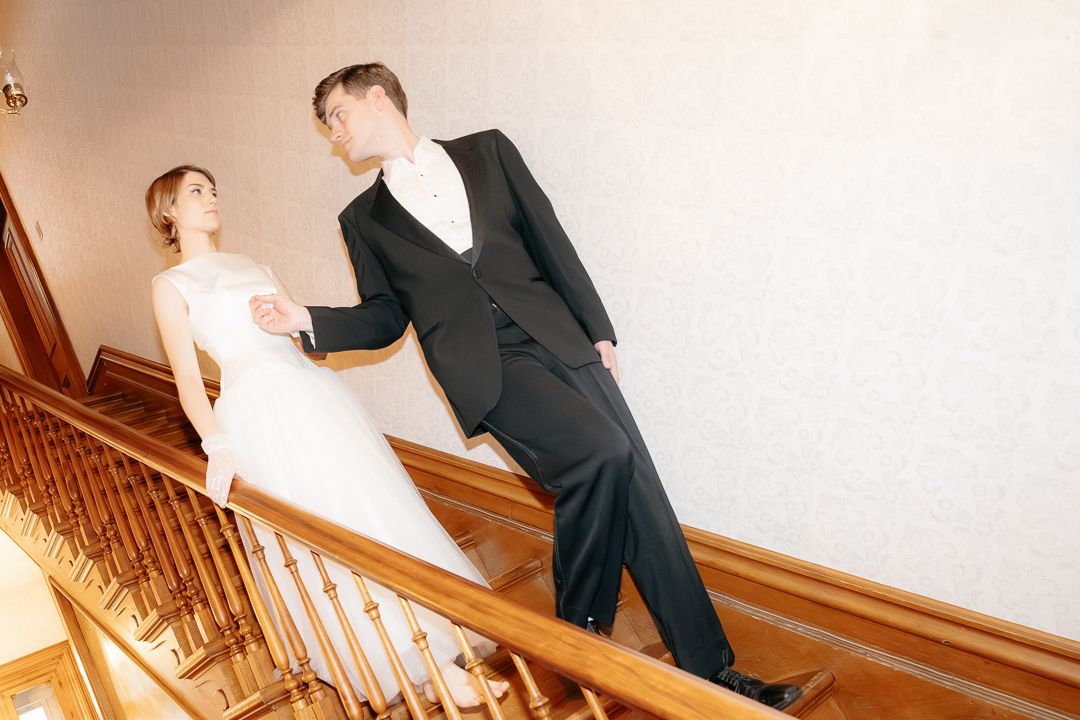 Bride and groom walking up elegant wooden staircase in wedding attire, white gown and black tuxedo.