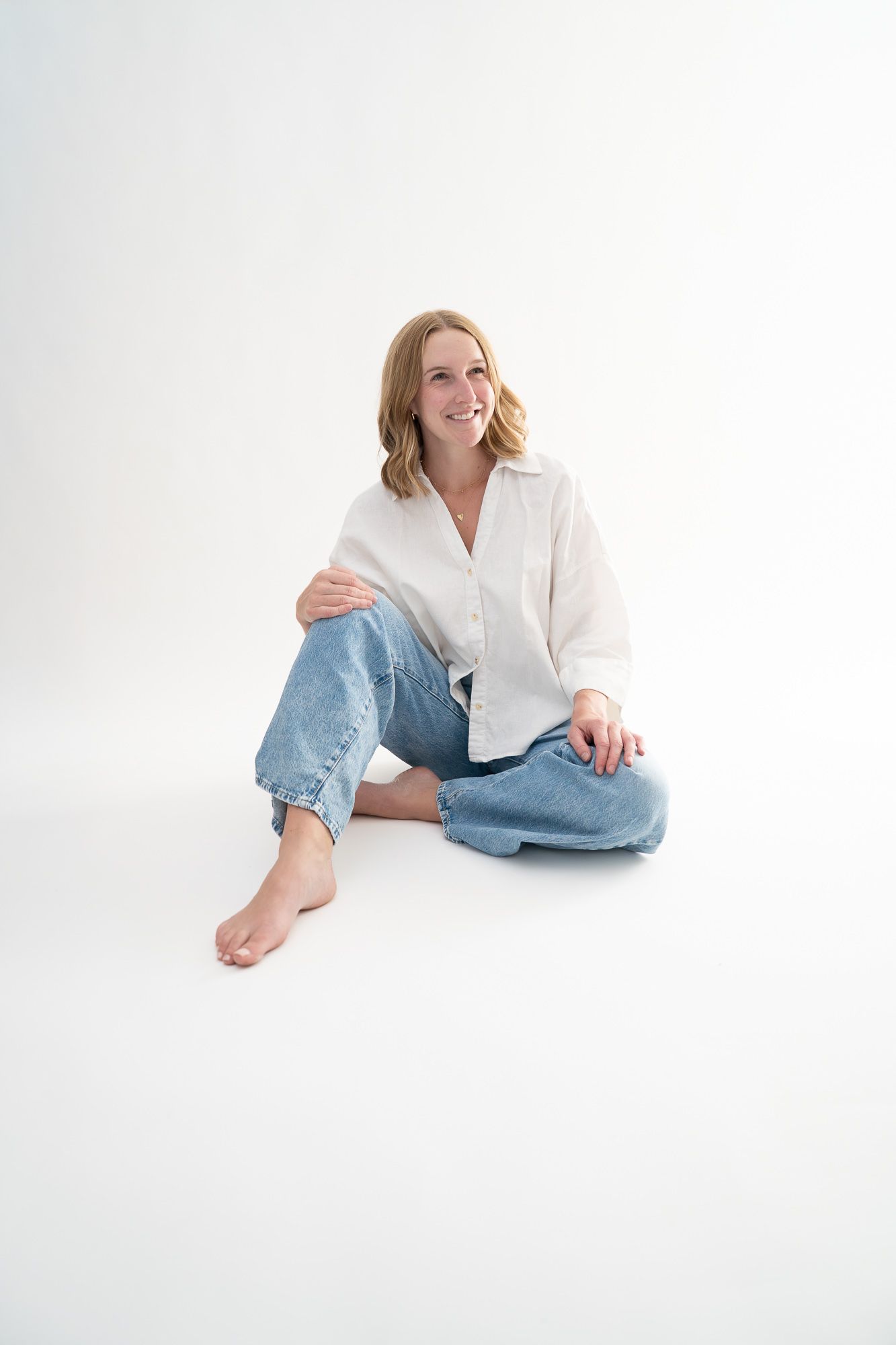Smiling blonde woman sitting cross-legged on white background wearing white linen shirt and blue jeans, barefoot.