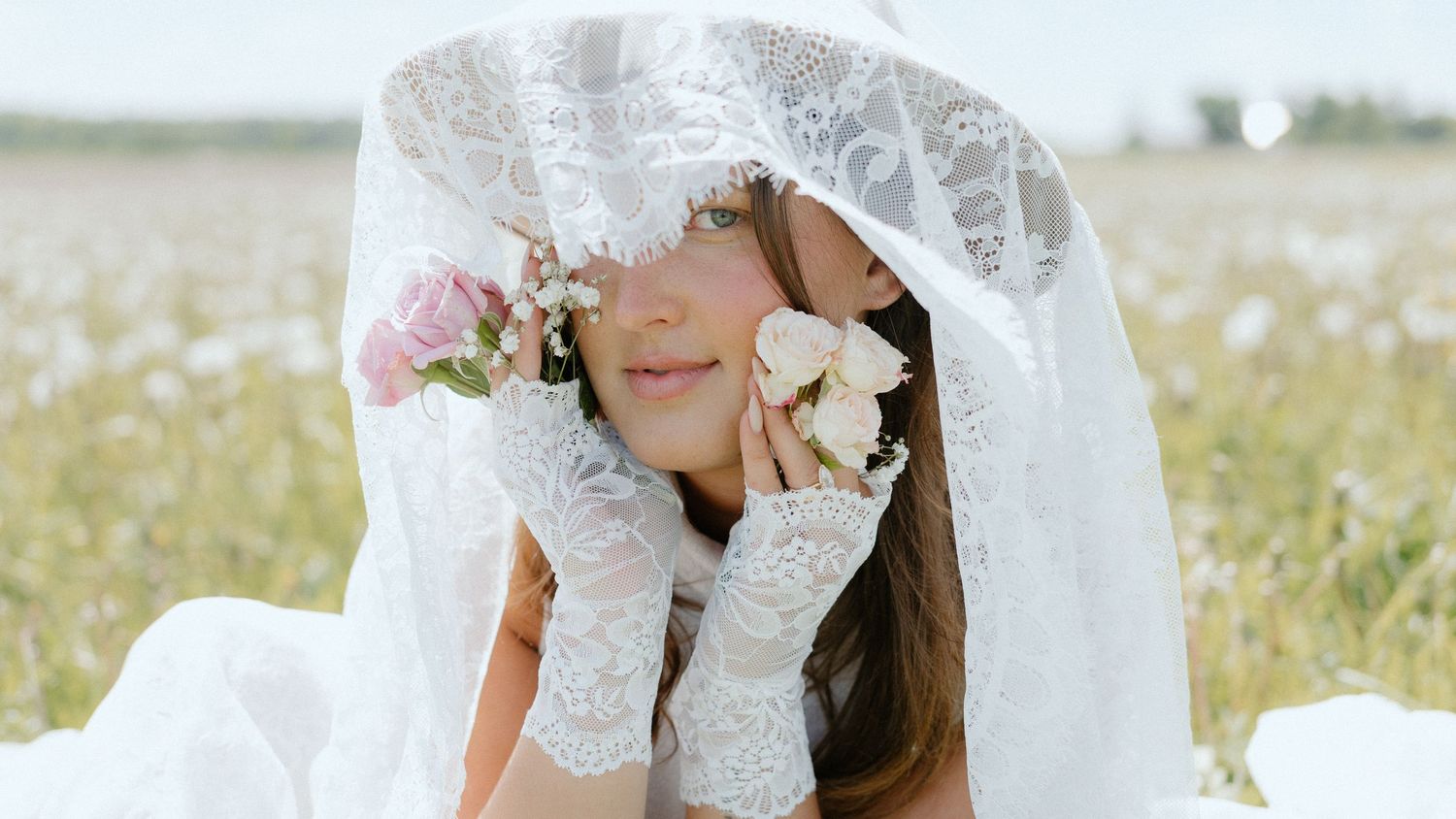 A bride in a white lace veil holds pink roses in a sunny flower field, wearing lace gloves in a dreamy outdoor setting.