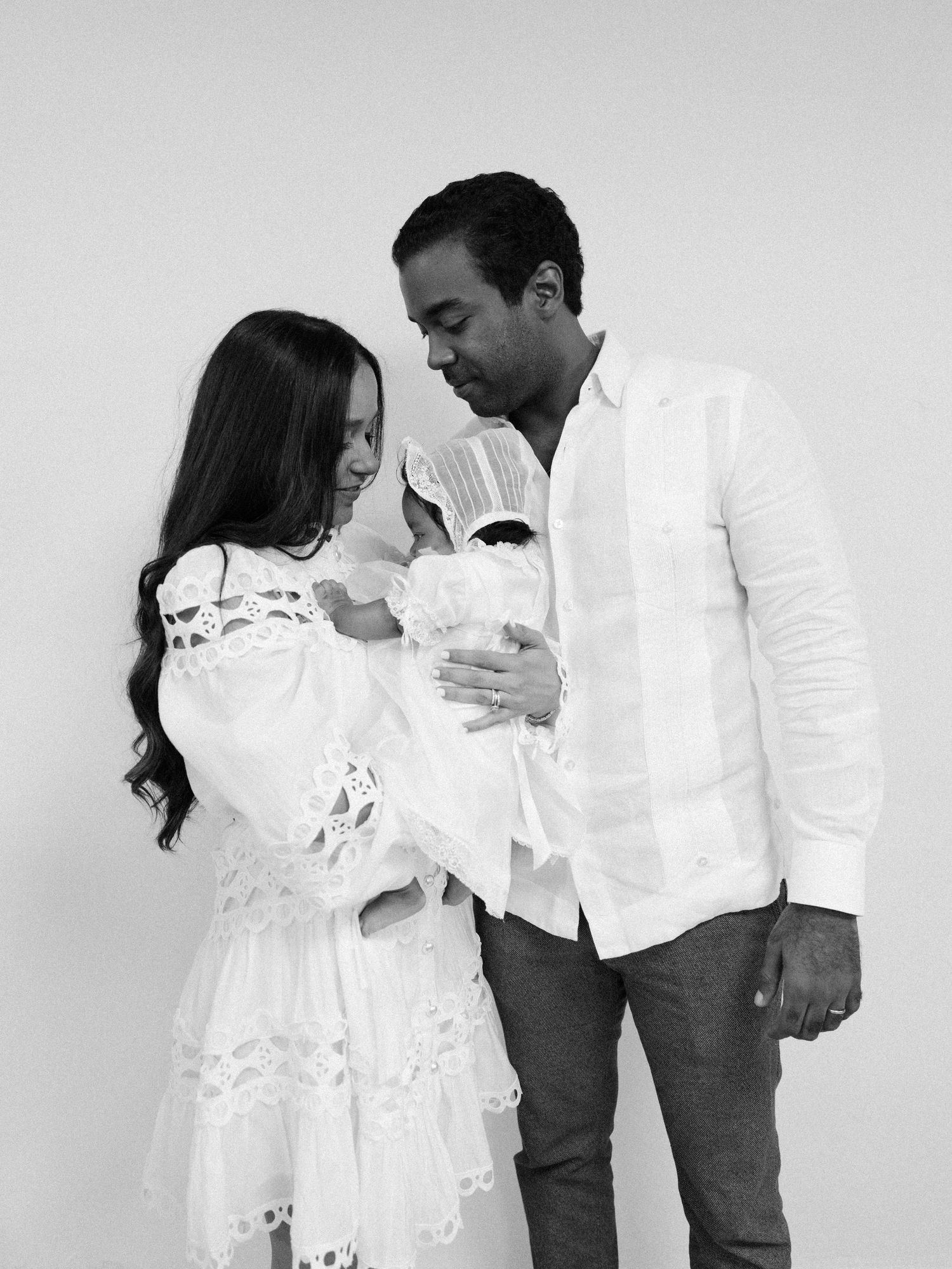 Black and white photo of a couple in white attire lovingly holding their newborn baby during a family portrait session.