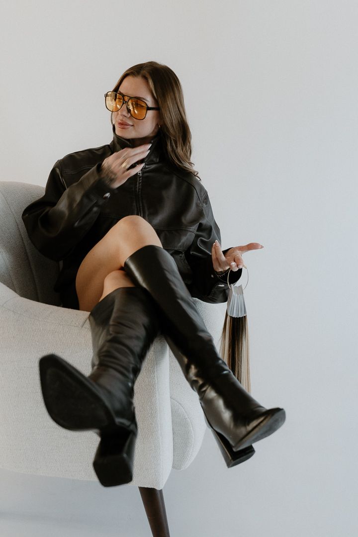 Woman in black leather jacket, knee-high boots, and amber sunglasses sitting confidently in a white armchair.