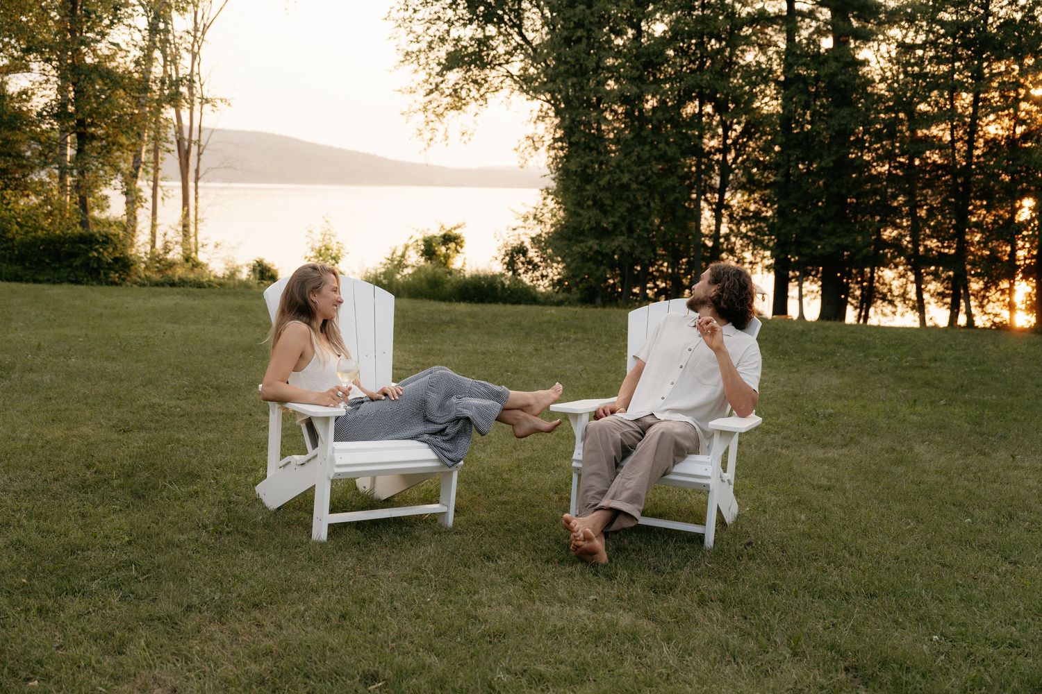 Couple relaxing in white Adirondack chairs on a lakeside lawn at sunset surrounded by trees.