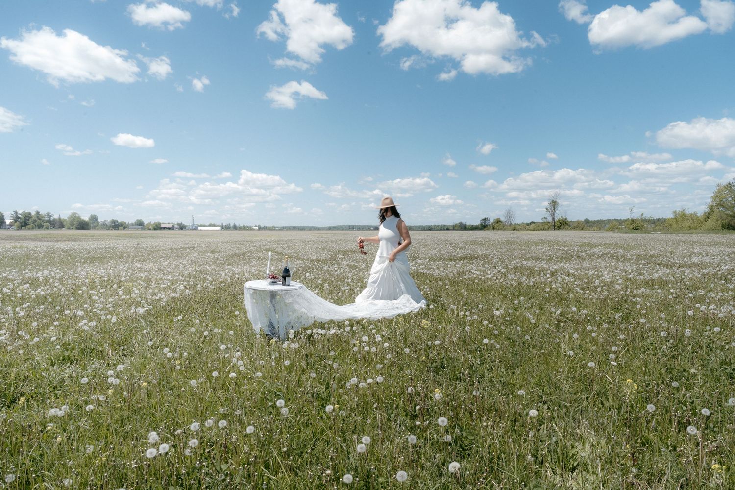Bride in white dress and hat stands in a vast wildflower meadow under a blue sky with fluffy clouds.