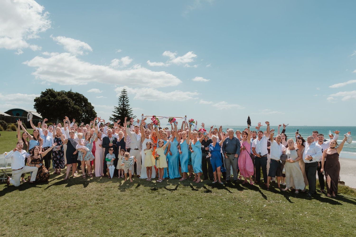 A large group of wedding guests in colorful attire pose together on a grassy hillside overlooking the ocean.