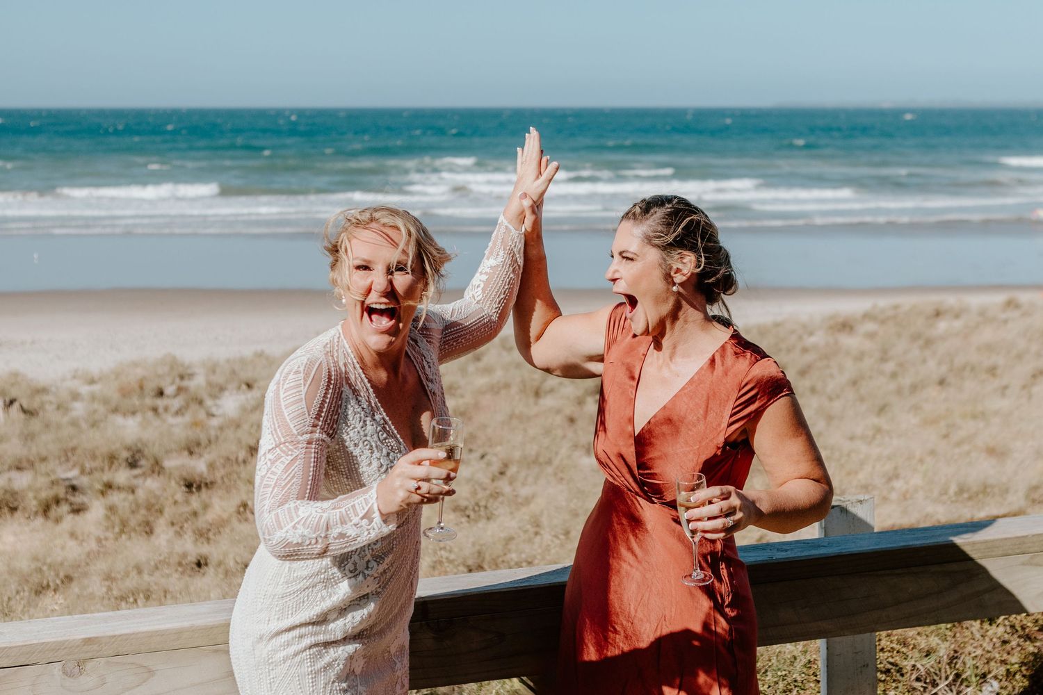 Two people high-fiving and laughing together at a sunny beach with ocean waves in the background.