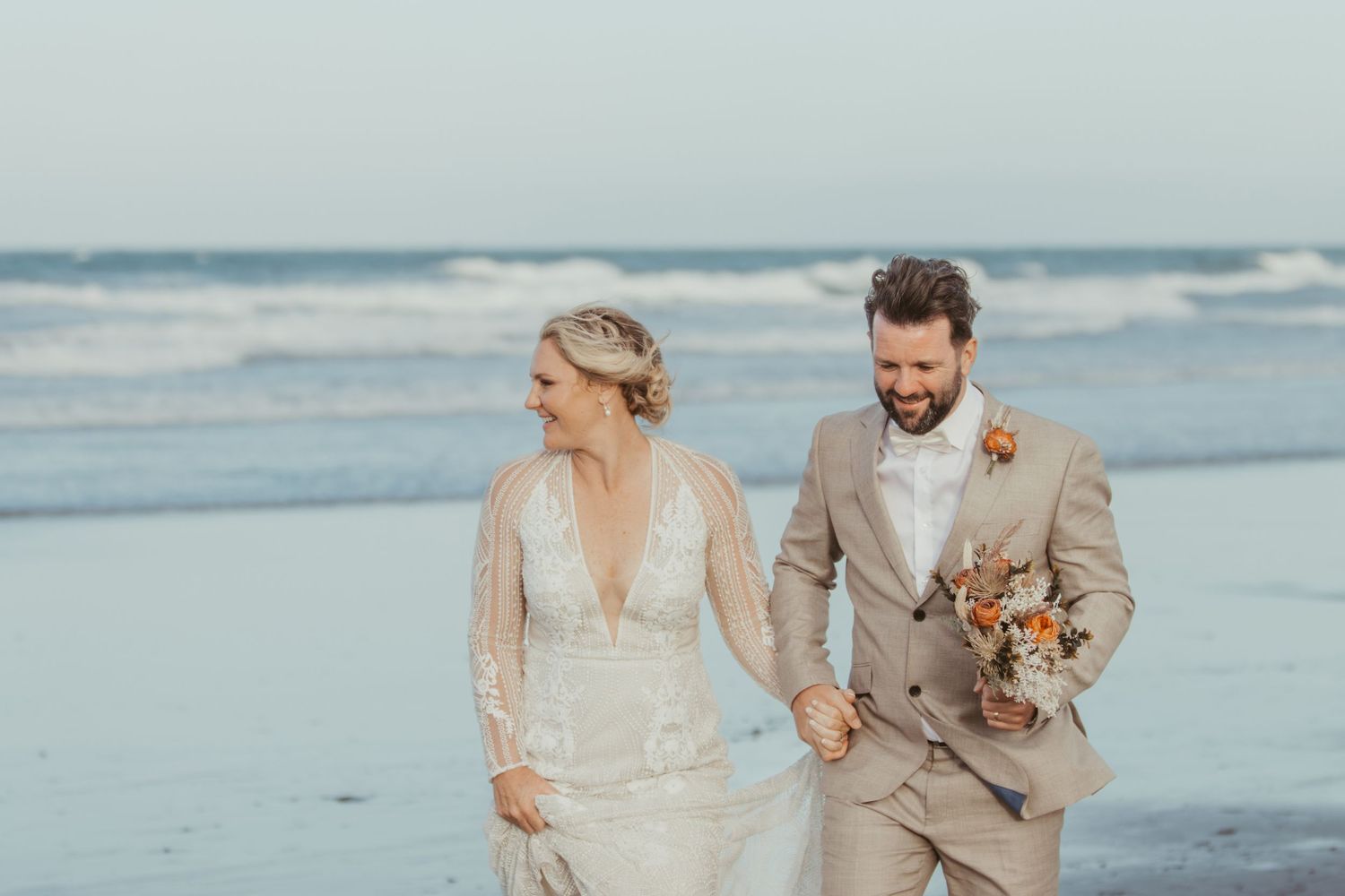 A couple walking along a misty beach at sunset wearing elegant wedding attire.