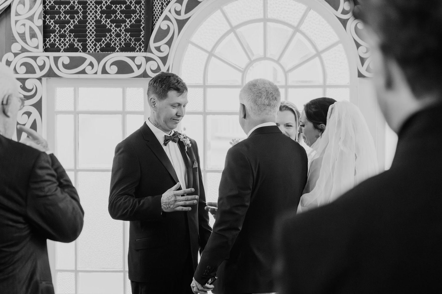 Groomsmen in black suits laugh together during a candid wedding moment in front of an elegant arched doorway.