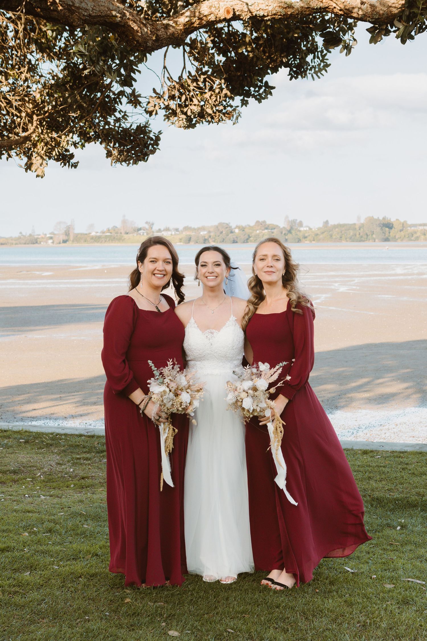 Bridesmaids in burgundy dresses pose with bride in white gown against coastal landscape backdrop.