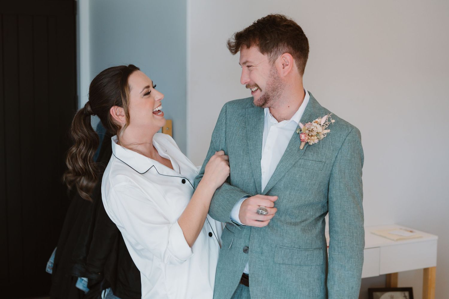 Professional stylist adjusts wedding boutonniere on groom's mint green suit jacket as they share a candid laugh.