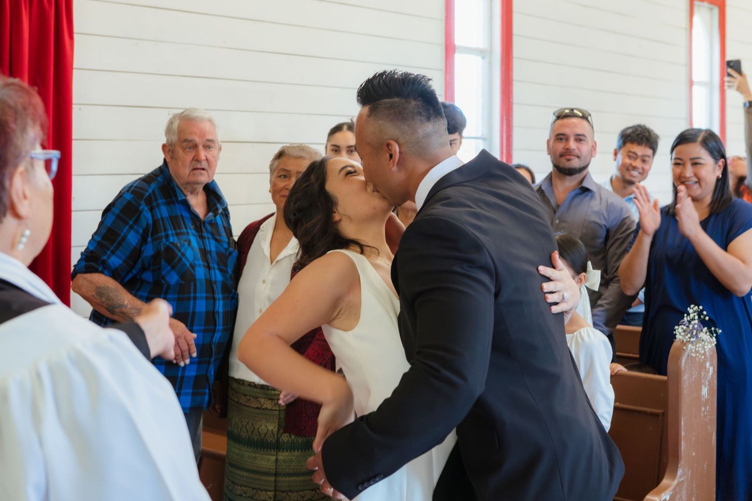 Two people share a romantic kiss during a wedding ceremony at a venue with red curtains and onlooking guests.
