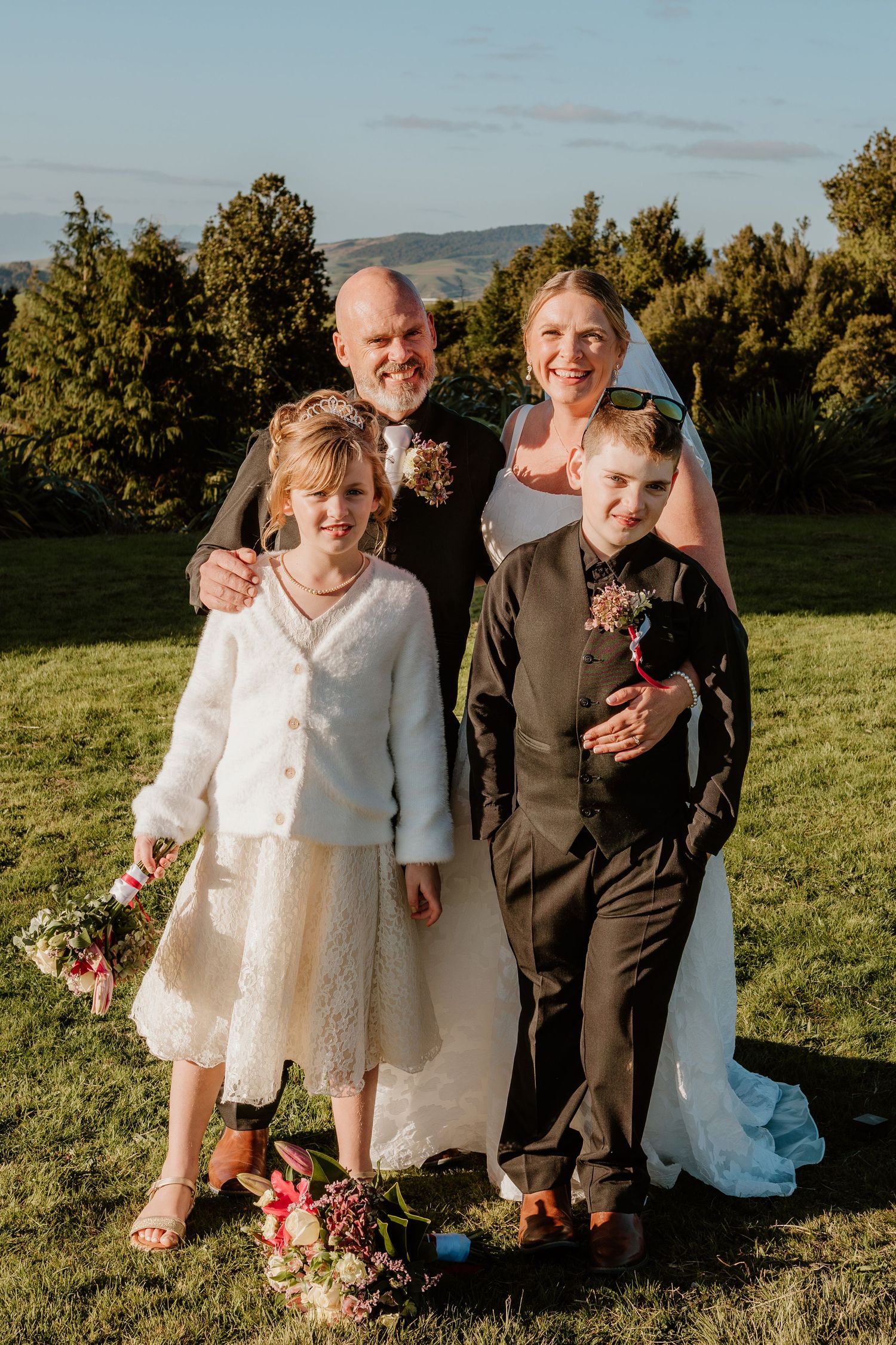 Happy wedding party poses together on grassy hilltop during golden hour with mountains and trees in background.