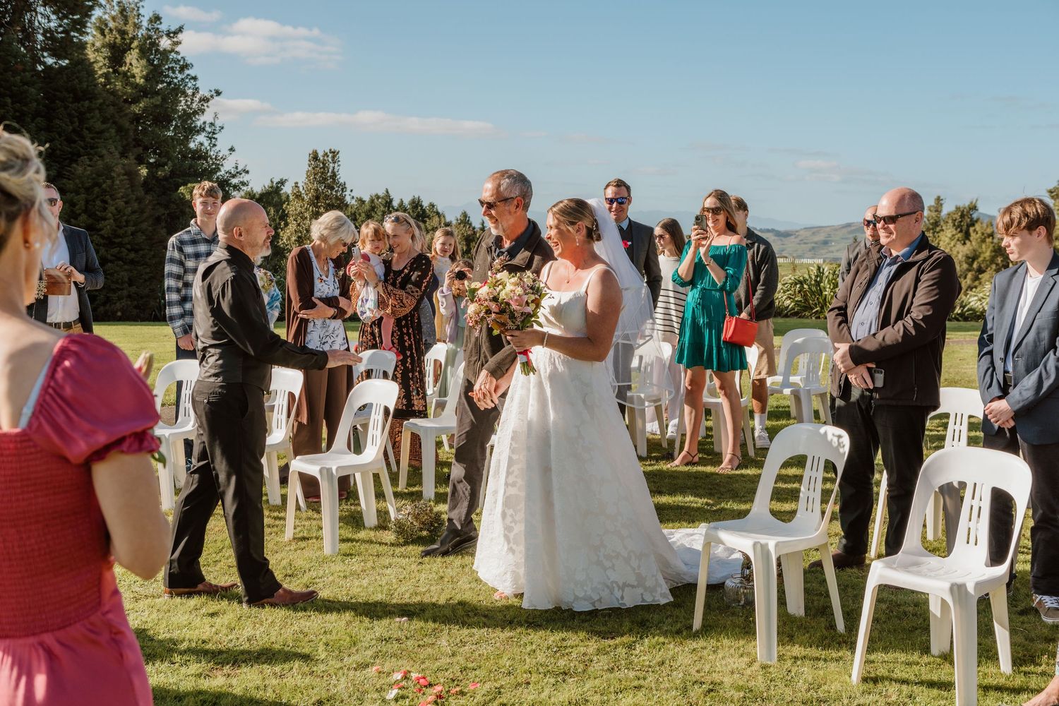 An outdoor wedding ceremony takes place on a grassy hilltop with white chairs and scenic mountain views in the background.