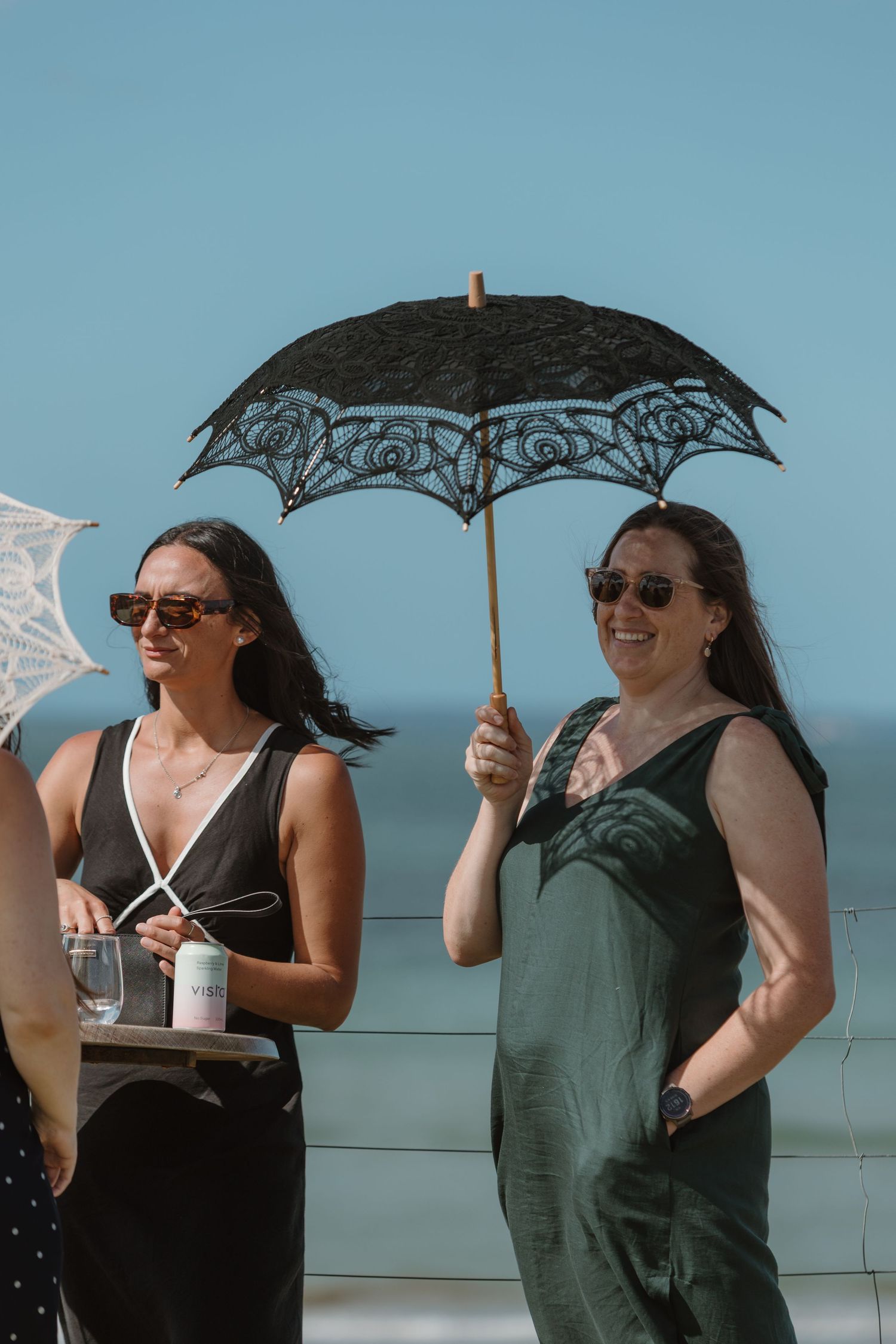 Two women at a beach event, one holding a black lace parasol umbrella, both wearing sunglasses on a sunny day.