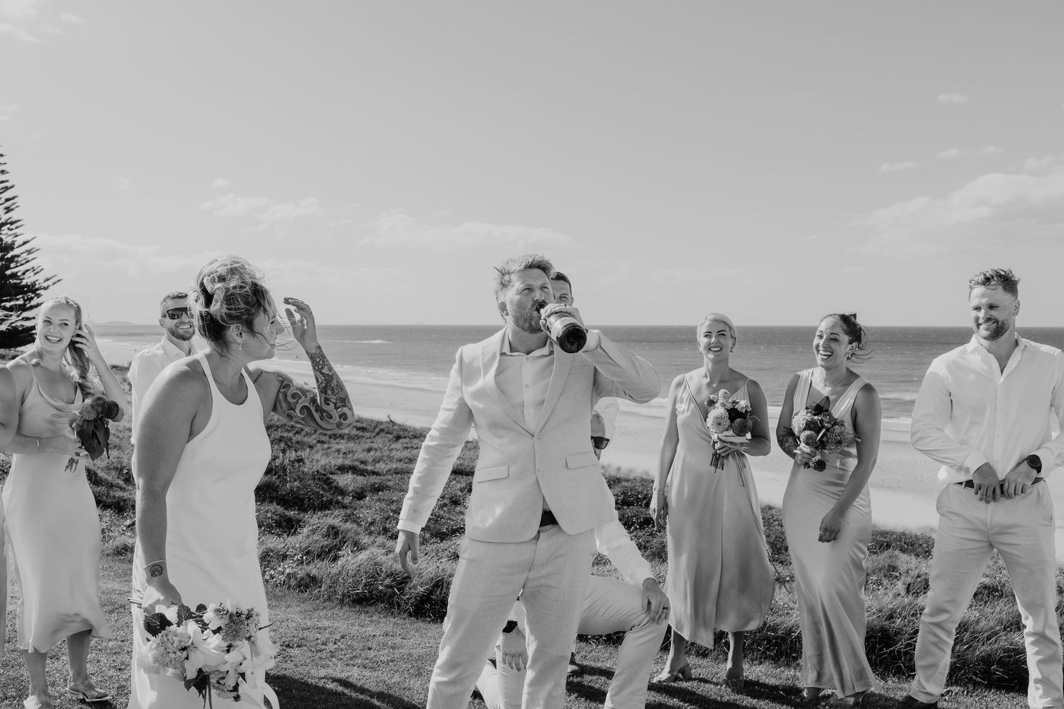 Black and white beach wedding party celebrating outdoors near ocean, bridal party in white and blue attire.