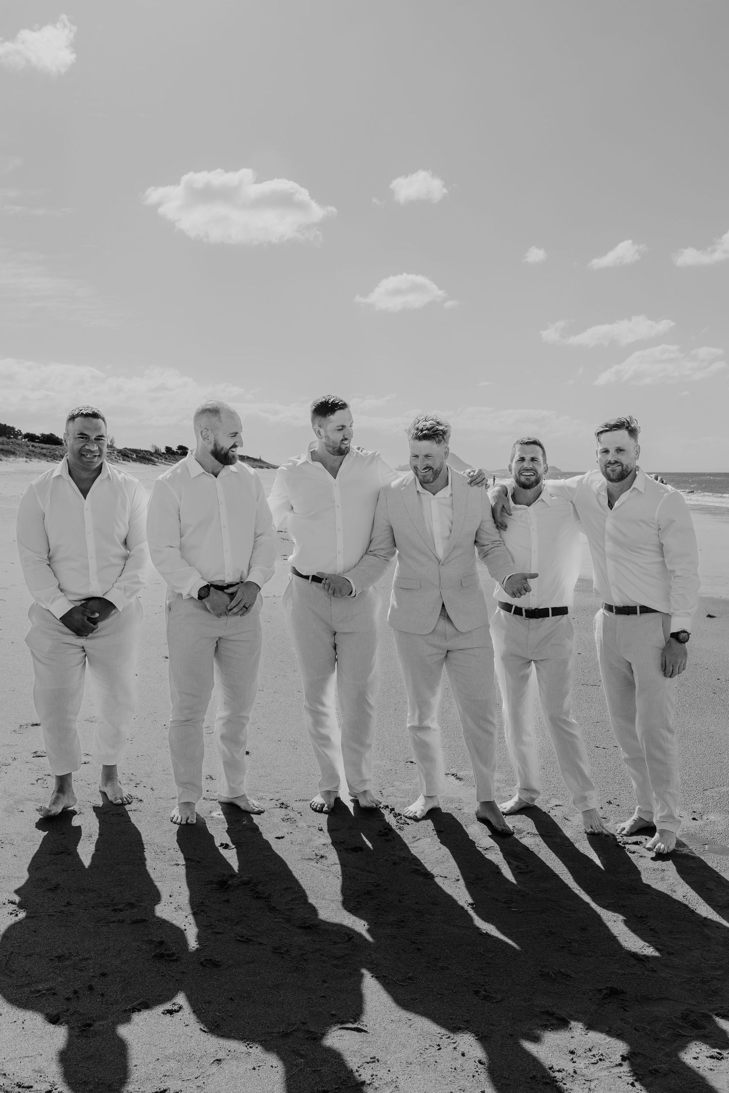 Six groomsmen in white linen suits pose together on a sunny beach in this elegant black and white wedding photo.