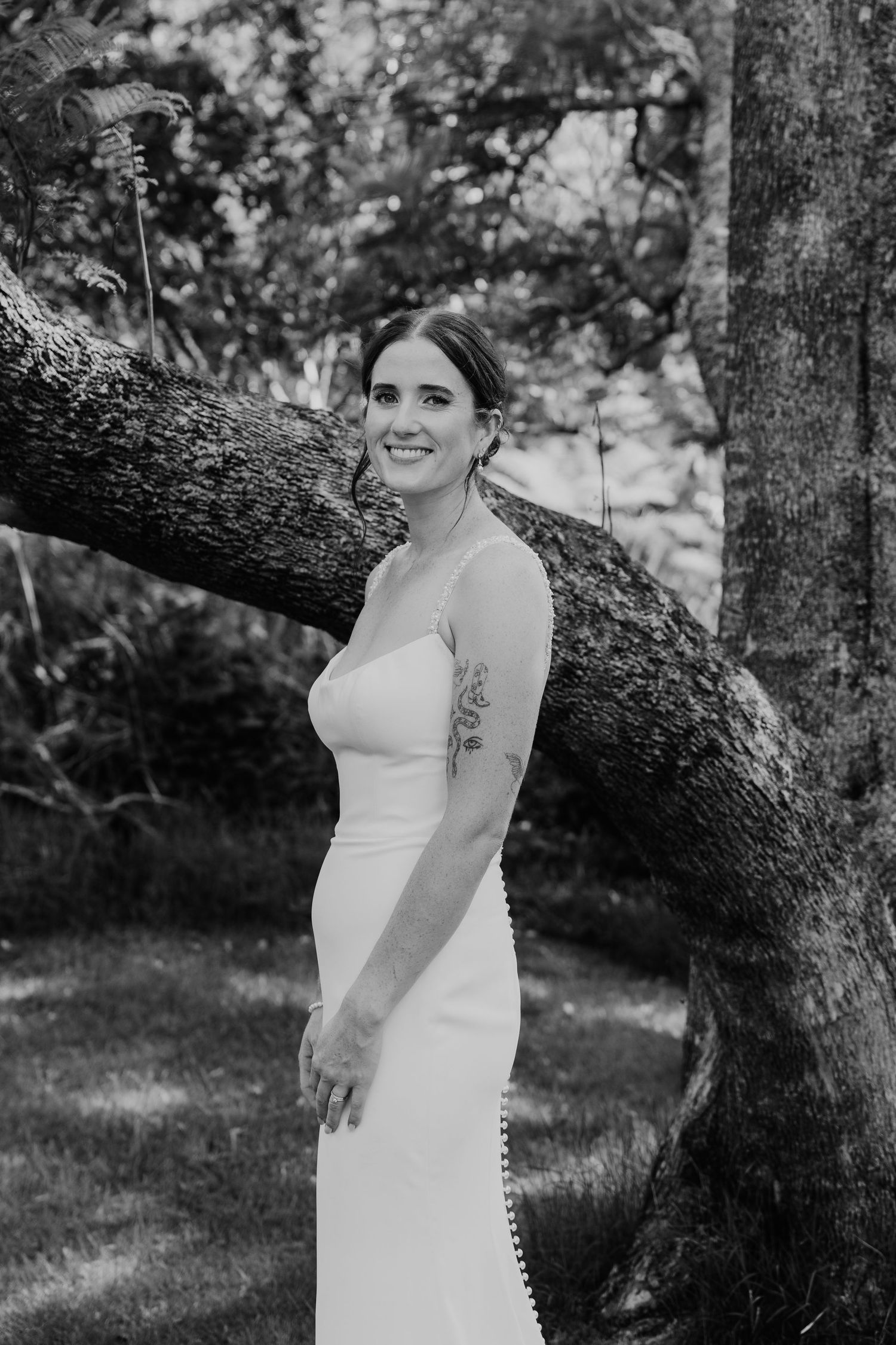 Smiling bride in white gown leans against a large tree trunk in a lush garden, black and white photo.