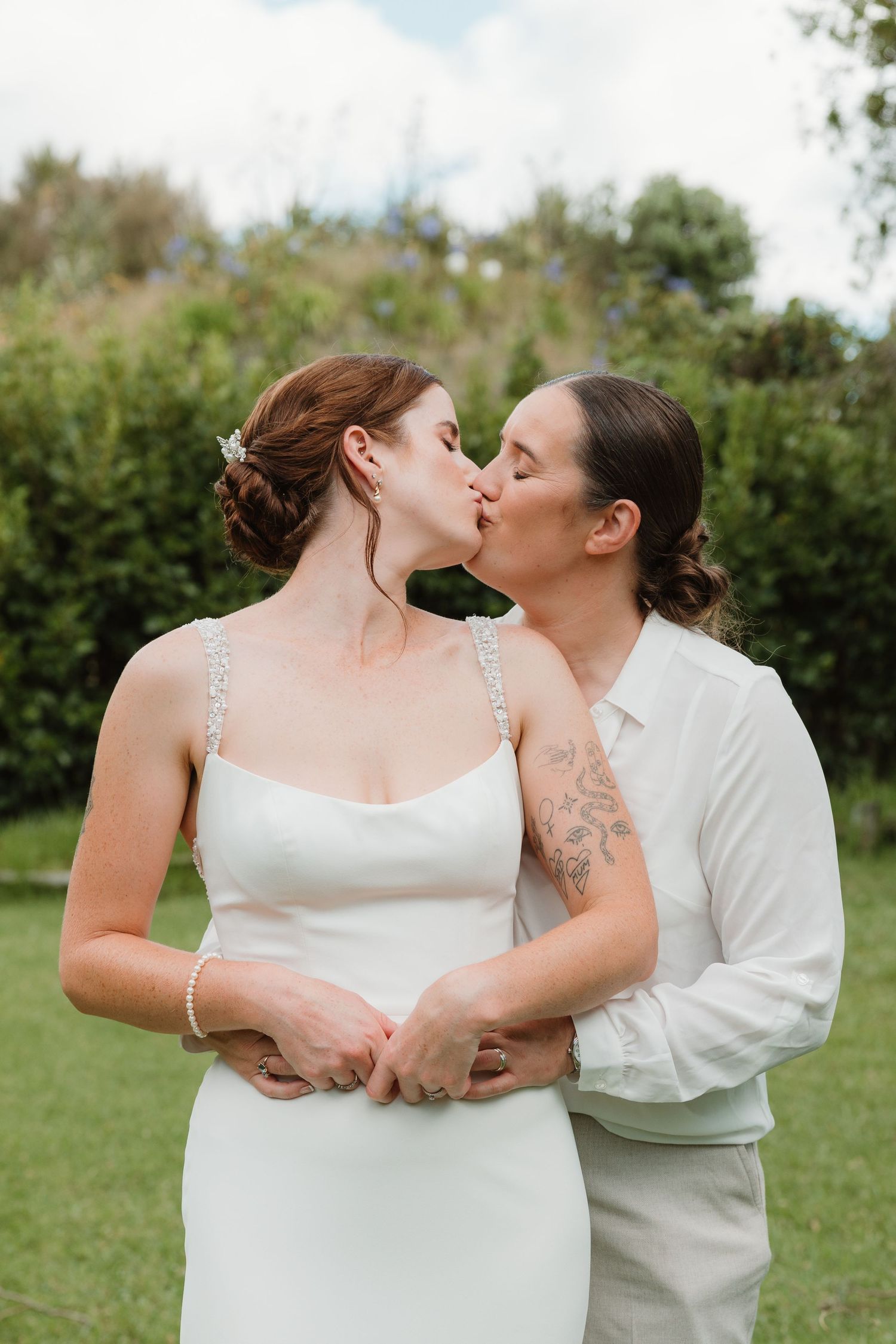 Two brides share a romantic kiss outdoors, one in a white gown and the other in a white shirt, surrounded by lush greenery.