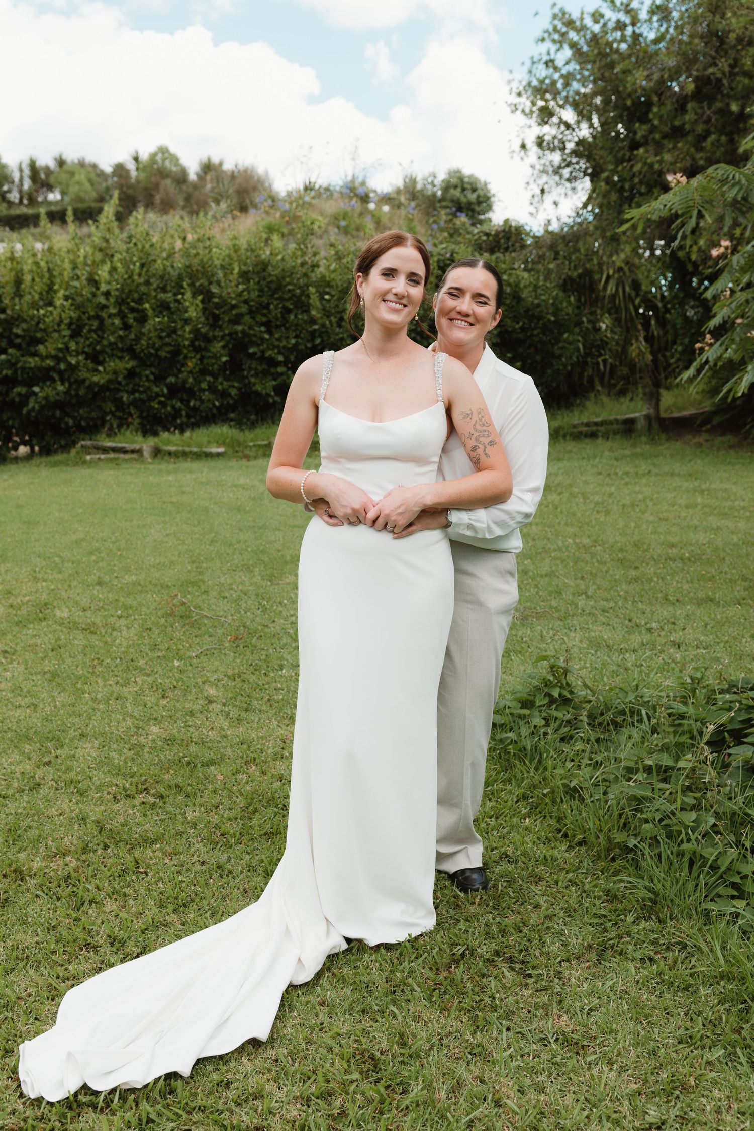 Two brides pose together outdoors on green grass, one in a white gown with train, the other in a white suit.