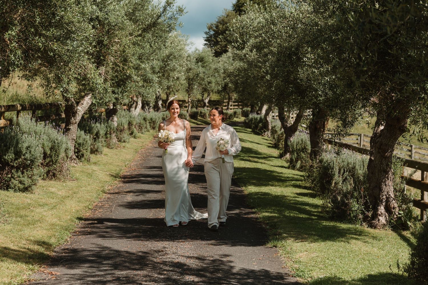 Two brides walk hand-in-hand down a tree-lined path surrounded by olive trees on their wedding day.