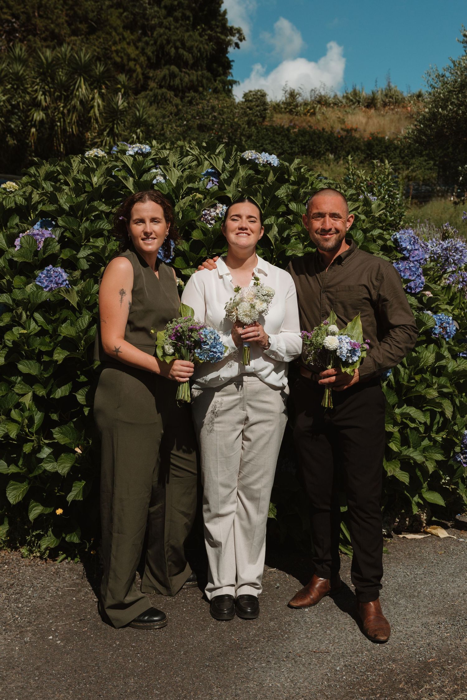 Three people holding blue hydrangea bouquets pose outdoors in front of lush green hydrangea bushes on a sunny day.