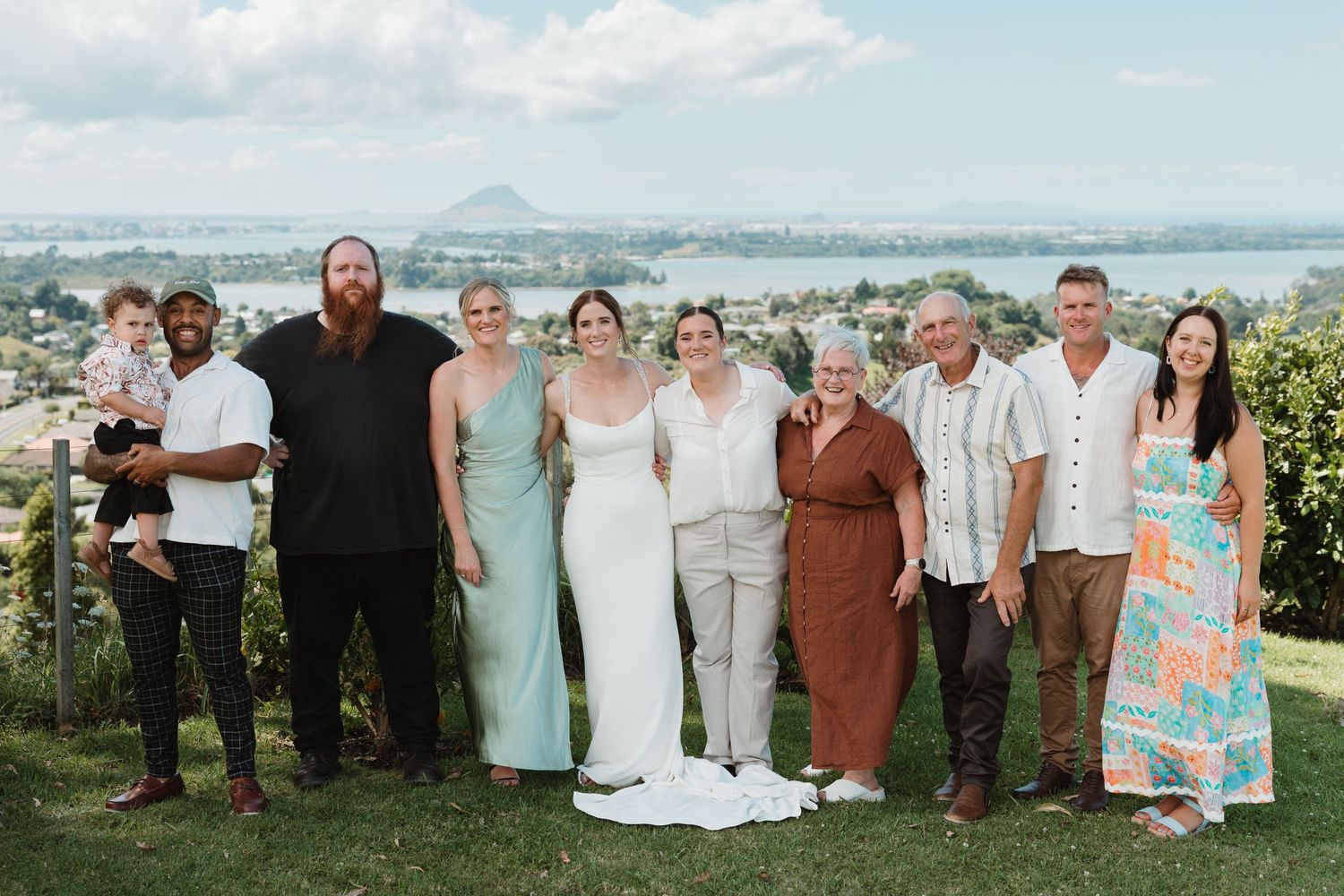 Wedding party posing outdoors with scenic water and mountain views in the background on a sunny day.