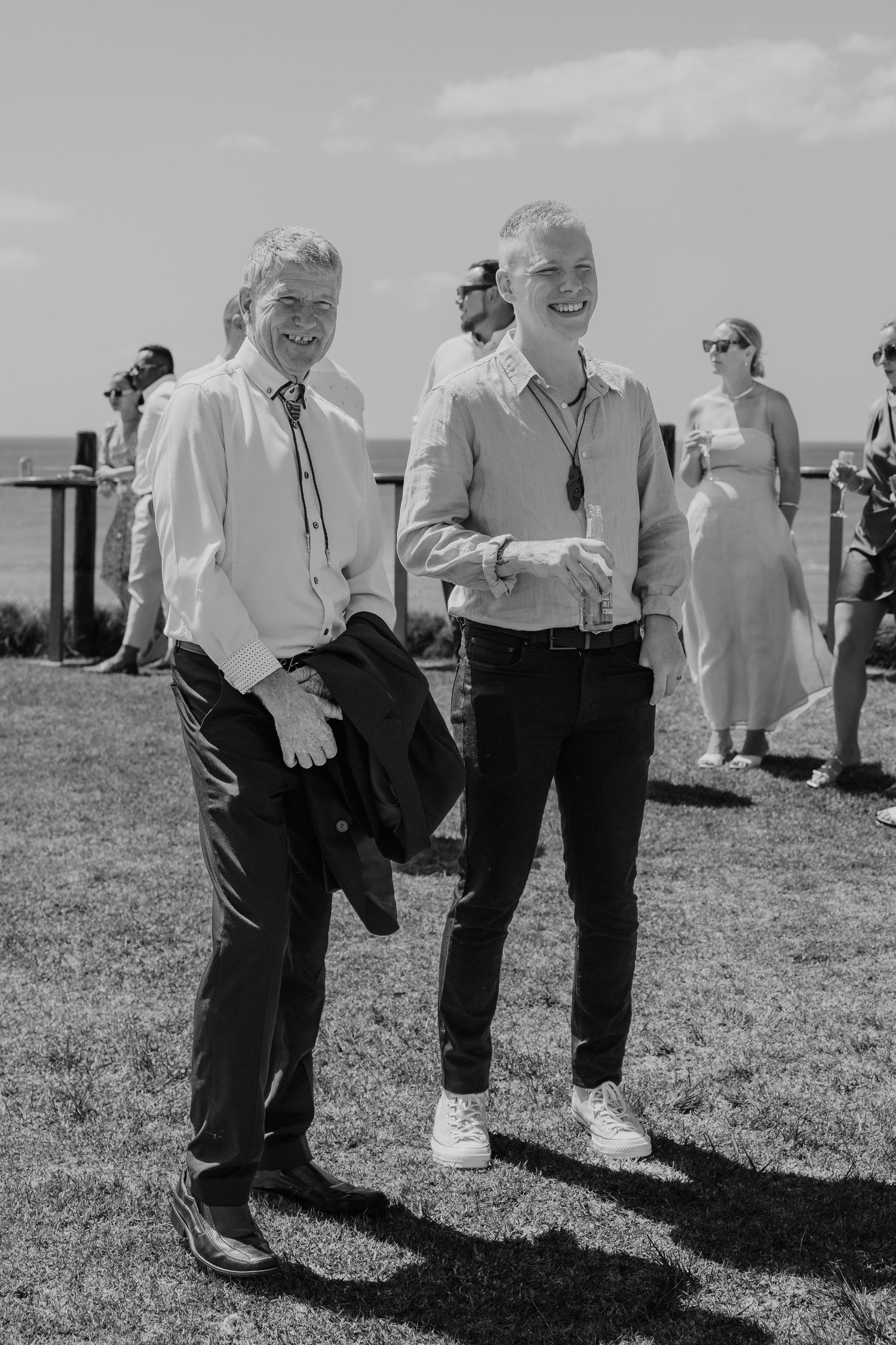 Two men in smart casual attire smile outdoors at a seaside event on a sunny day.