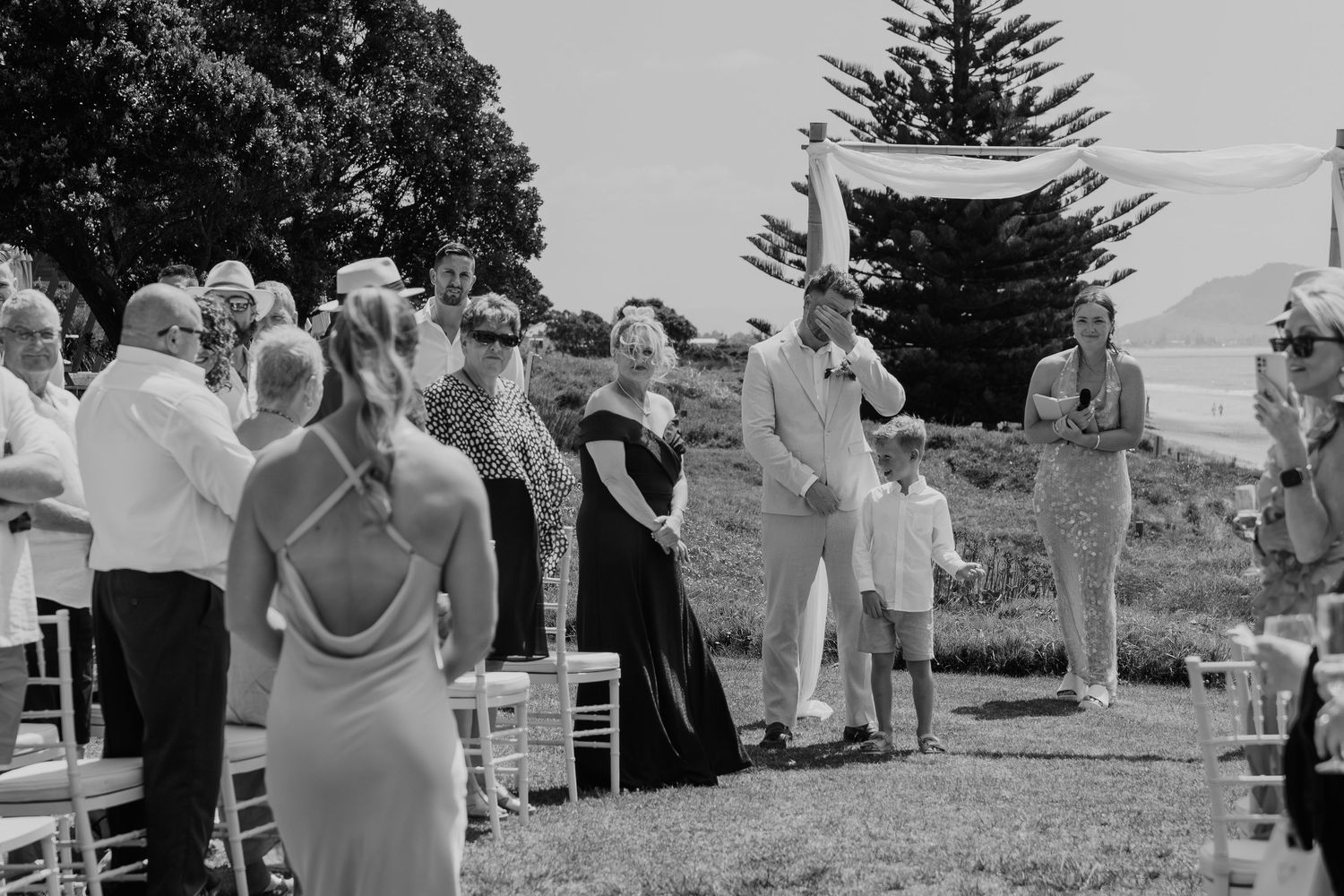 Black and white outdoor wedding ceremony with guests seated and standing on a grassy coastal lawn beneath a large tree.