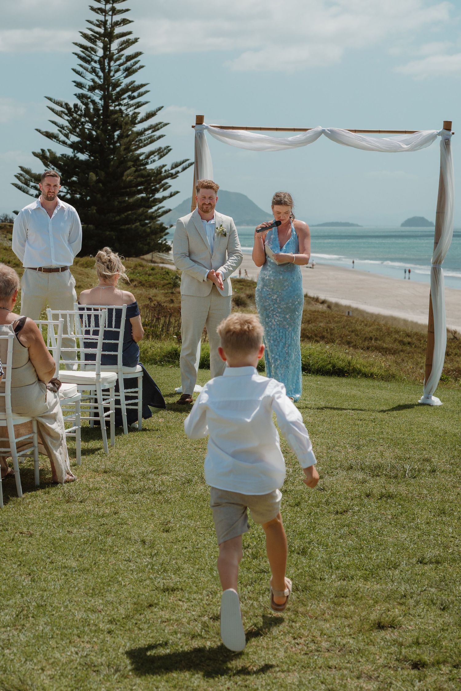 Young boy running toward wedding couple at outdoor beach ceremony with white arch and ocean backdrop.