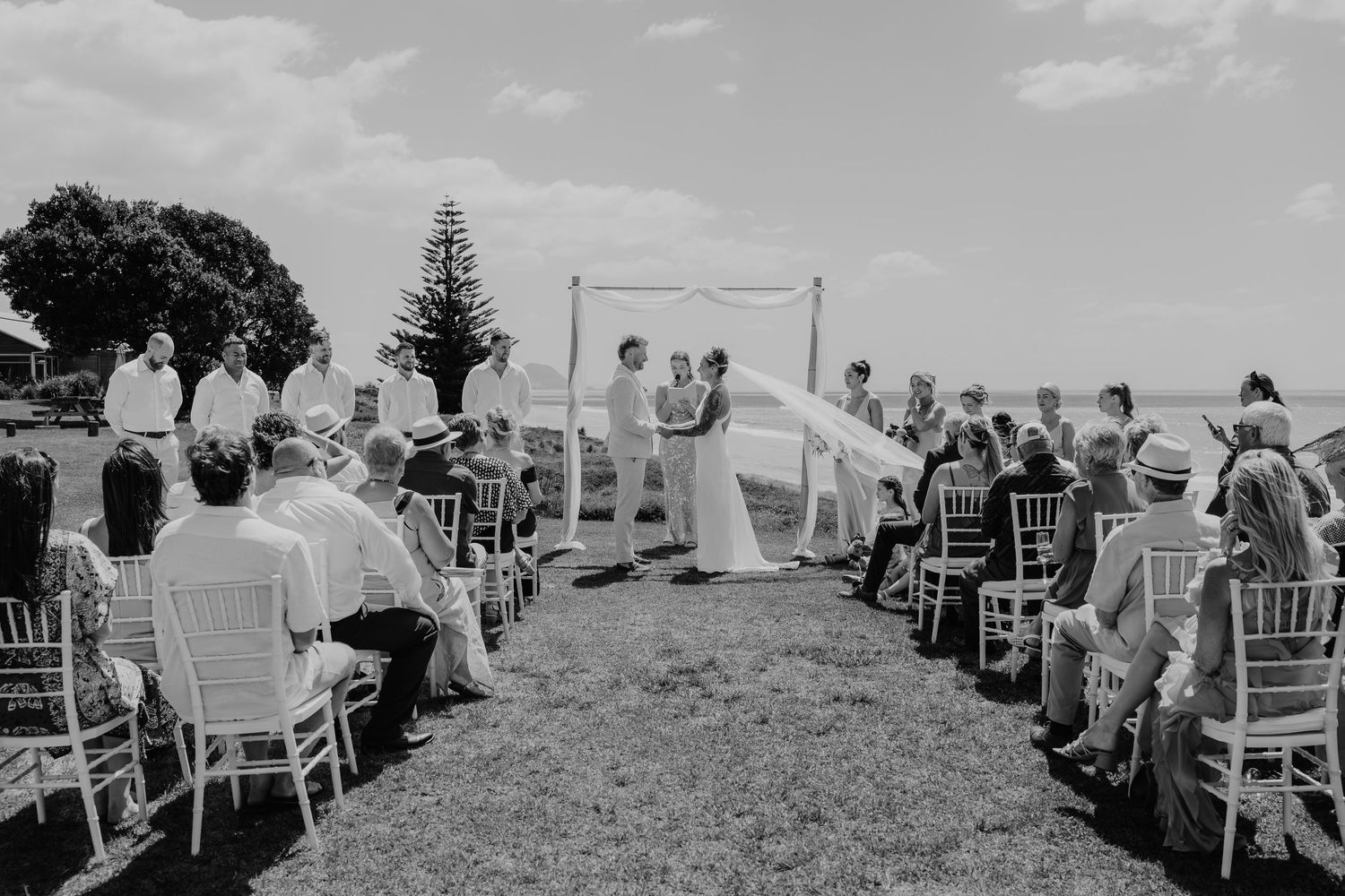 Black and white outdoor wedding ceremony with couple at altar, guests seated on white chairs on a grassy coastal lawn.