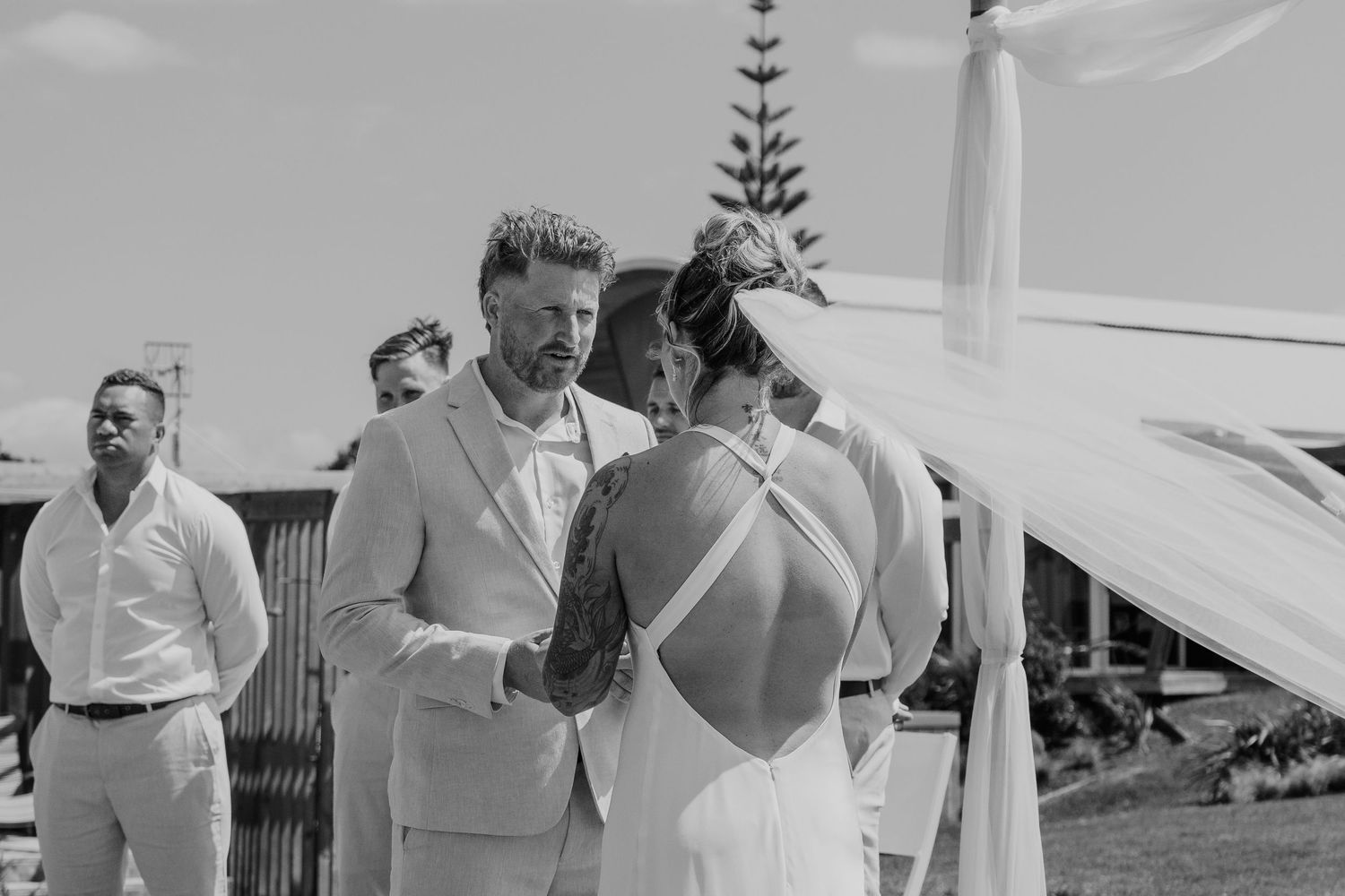 Black and white photo of a couple exchanging vows at an outdoor wedding ceremony with guests in the background.