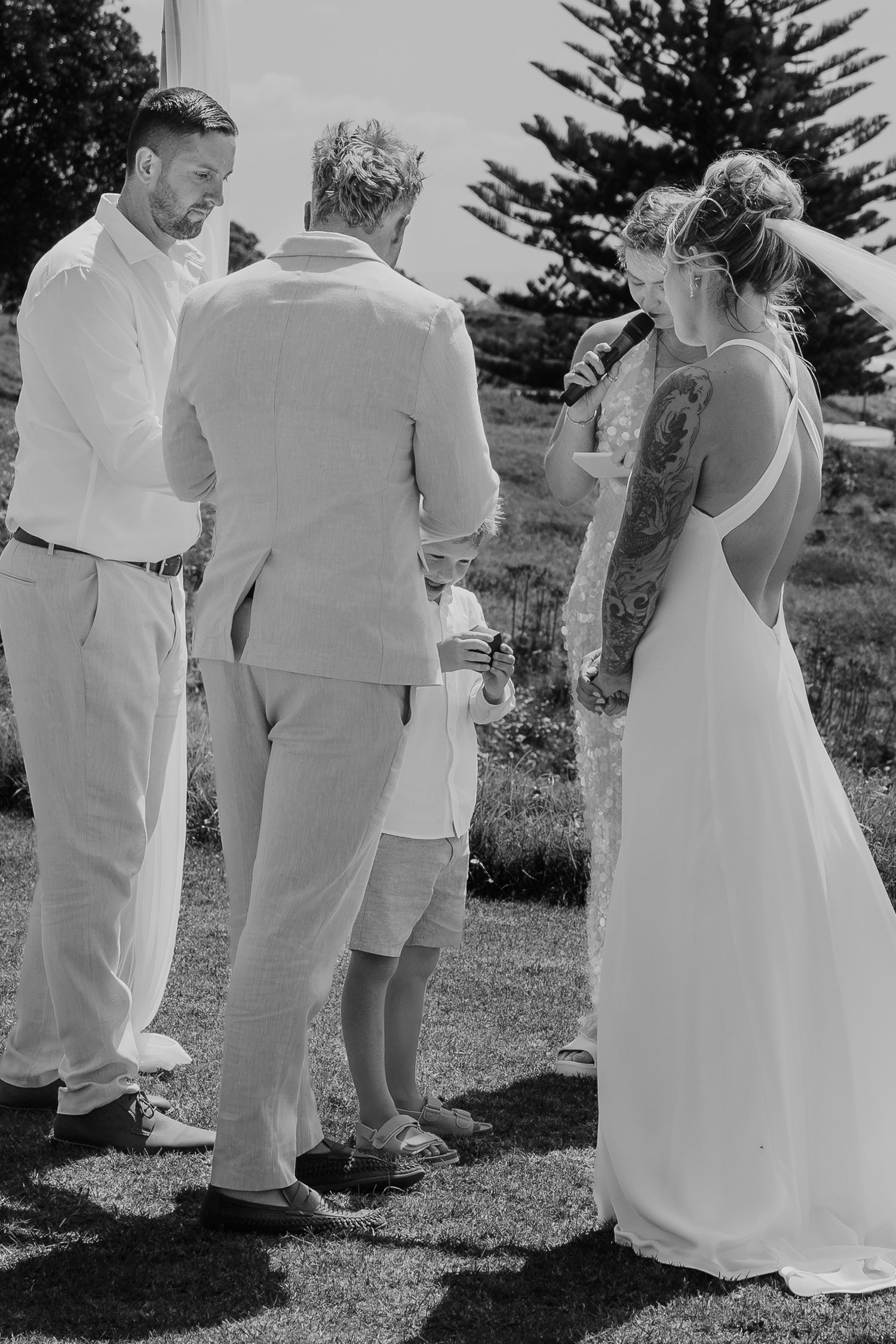 Black and white outdoor wedding ceremony with bride in backless gown, groom, and officiant exchanging vows.
