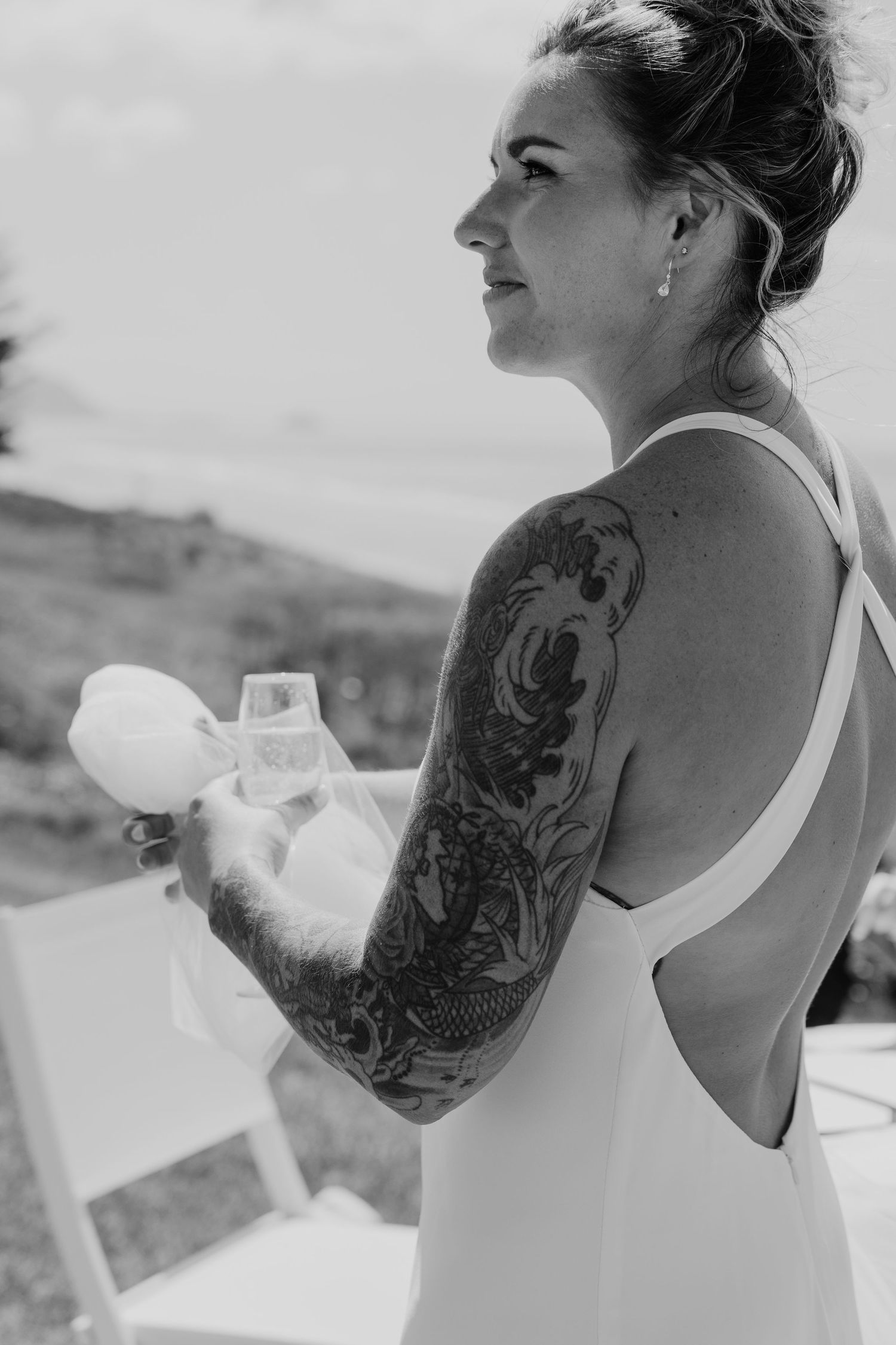 Black and white photo of a bride with a full sleeve tattoo in a backless wedding dress holding a champagne glass outdoors.