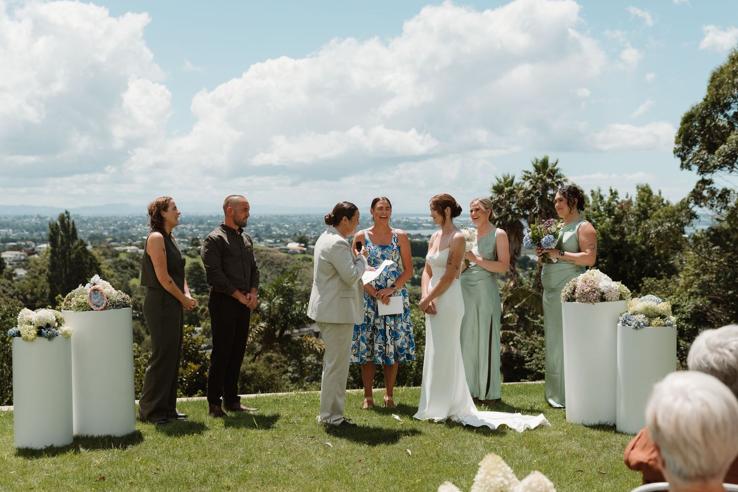 Outdoor wedding ceremony on a hilltop with bride, groom, officiant, and guests surrounded by lush greenery and blue skies.