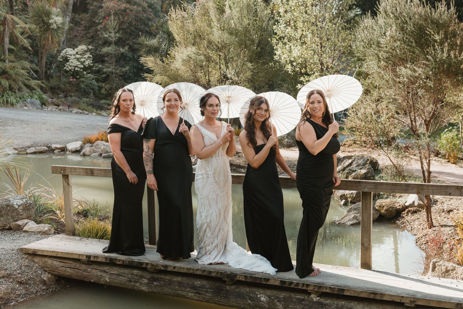 Wedding party in black dresses poses together by rustic wooden boat near waterfront.