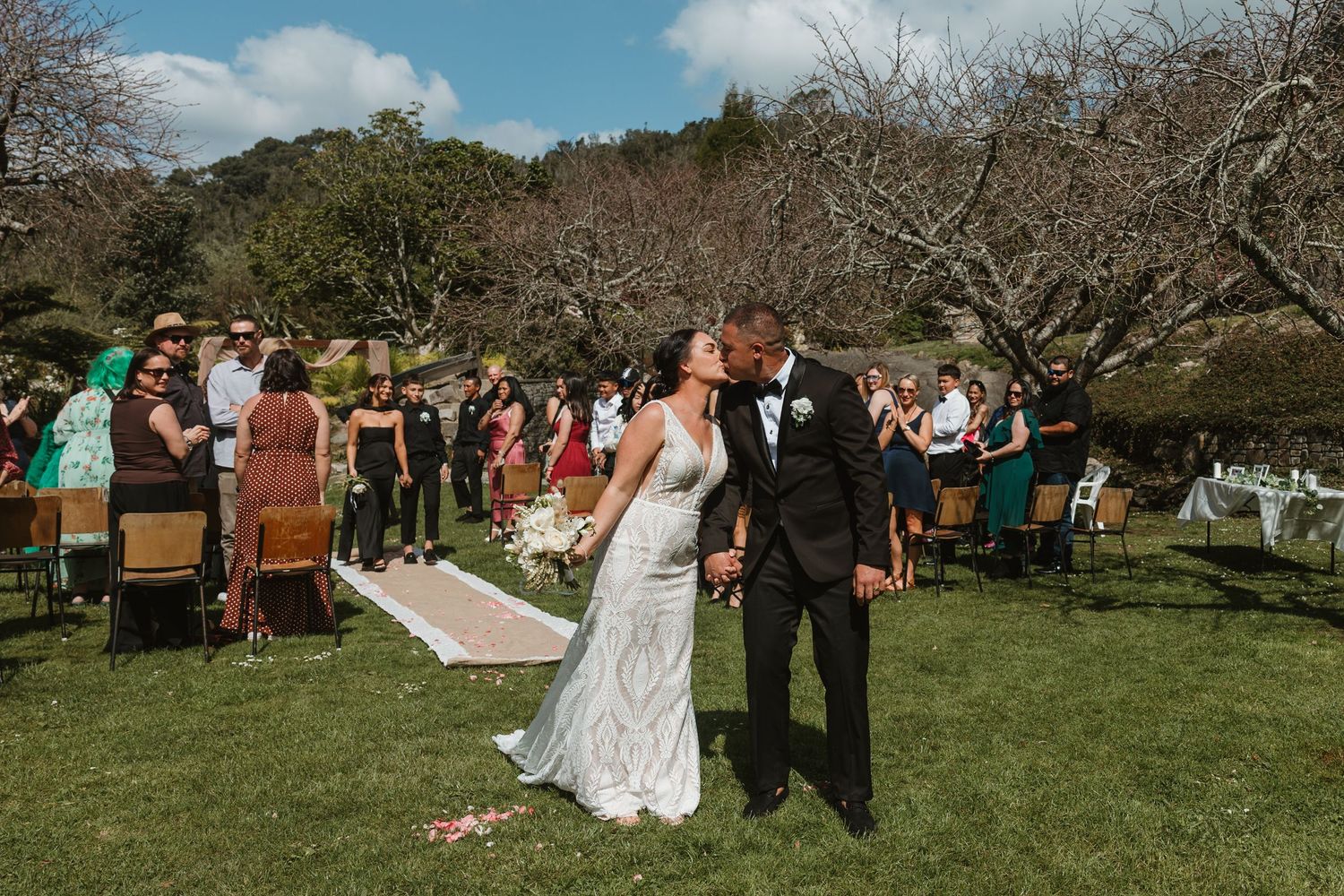 Outdoor wedding ceremony takes place in a scenic orchard setting with wooden chairs and natural decor.