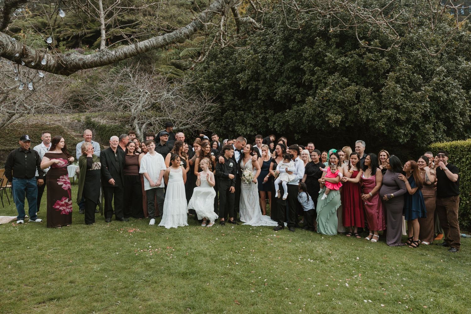 Large wedding party poses outdoors in formal attire for group photos on a green lawn near stone buildings.