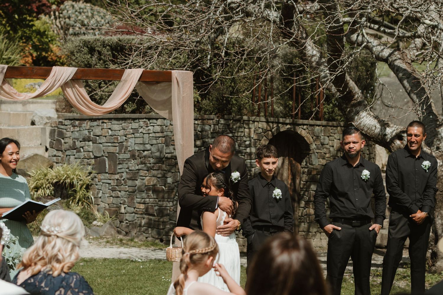 A wedding party celebrates outdoors in front of a rustic stone wall on a sunny day.