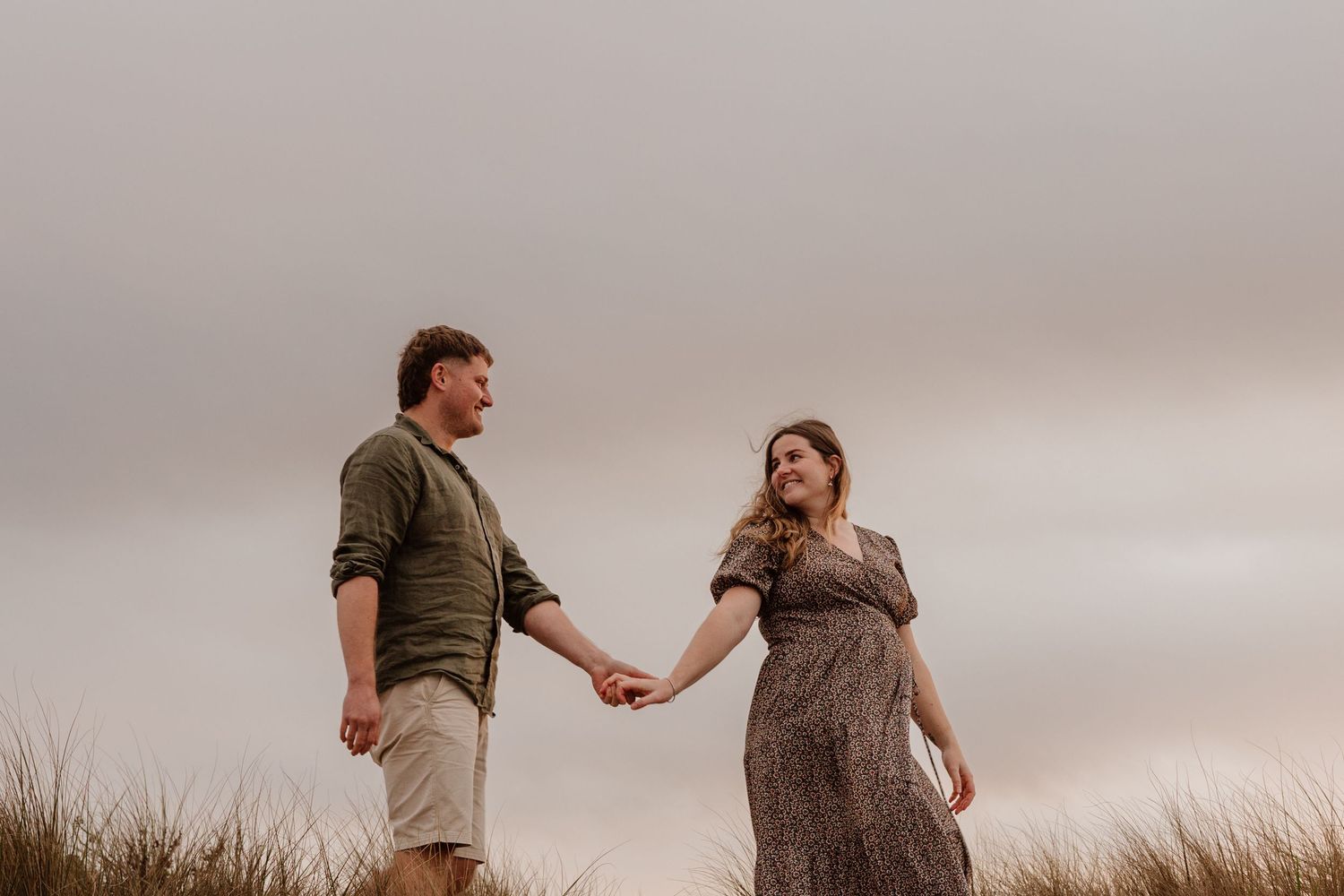 A couple walks through tall grass during sunset holding hands while wearing casual clothing.