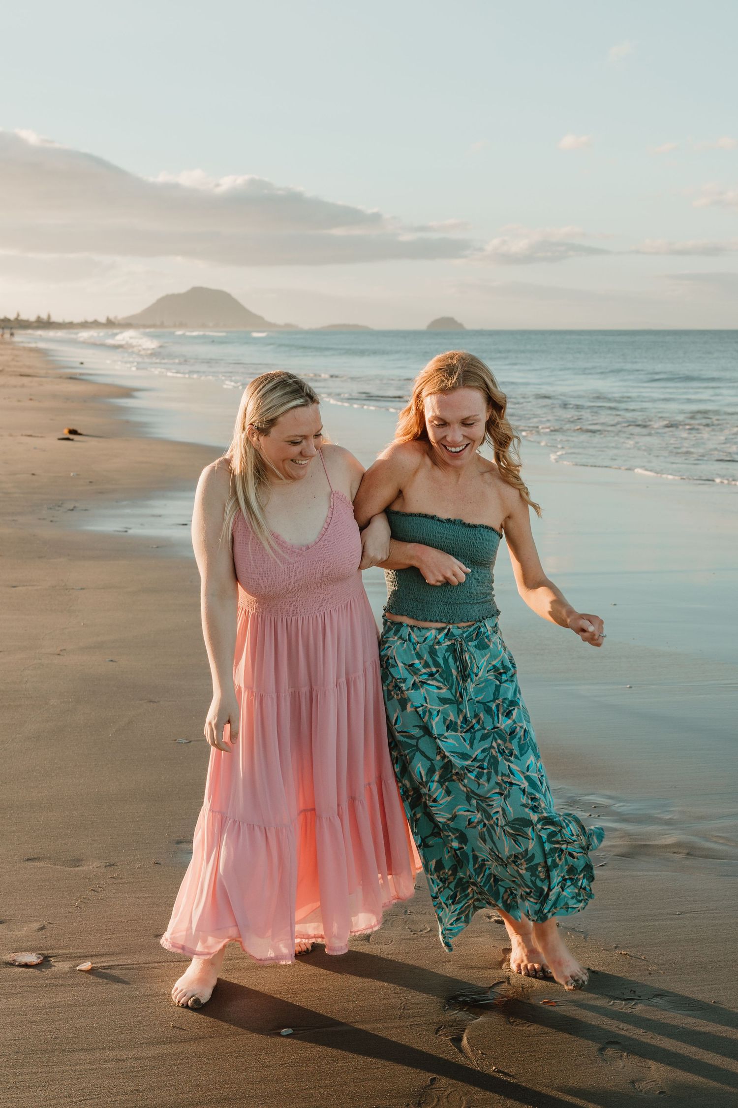 Two women in colorful summer dresses laughing and walking barefoot on a sandy beach at golden hour.