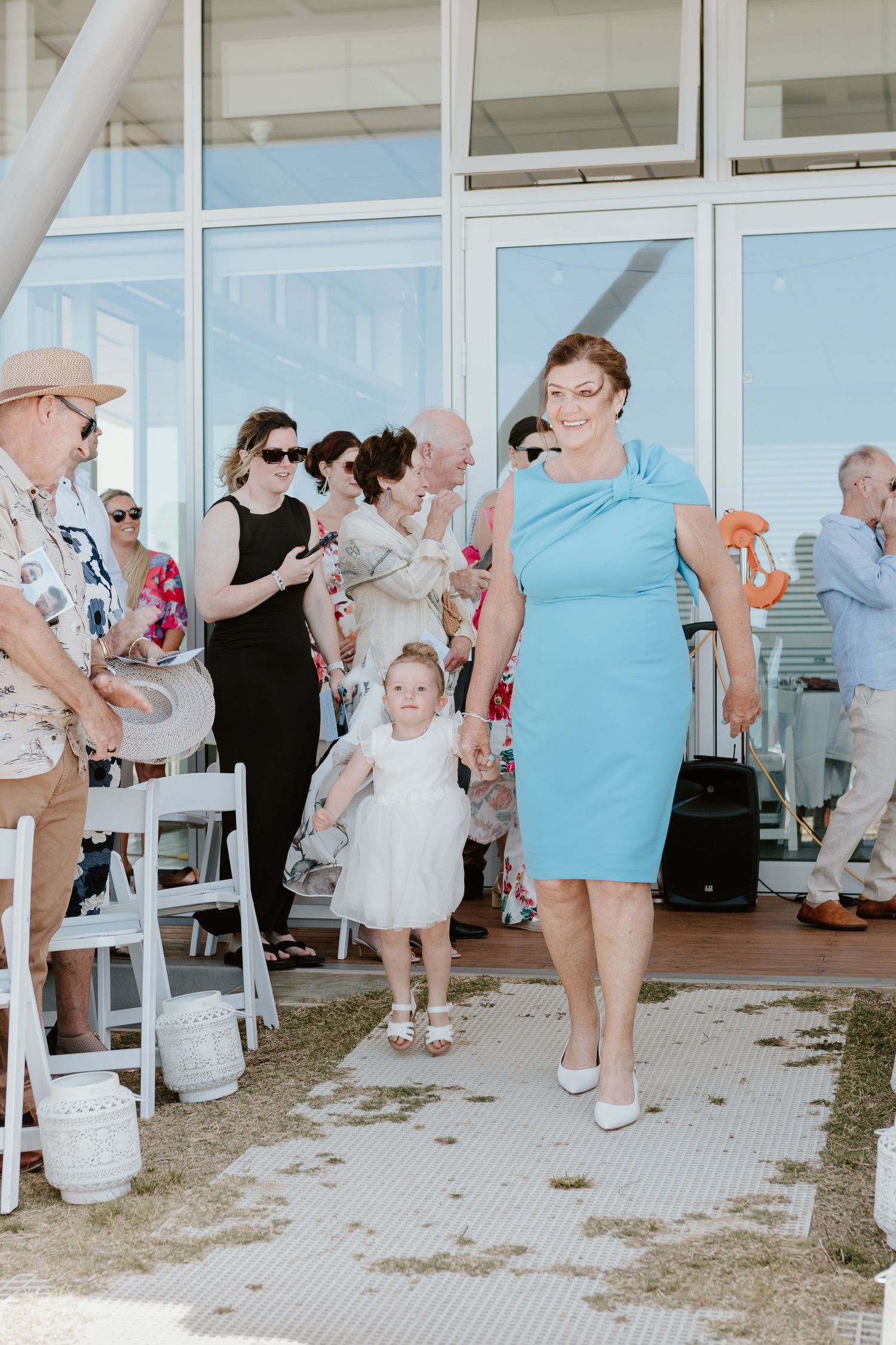 Wedding celebration with guests and child in white dress at coastal venue. Papamoa Surf Lifesaving Club Wedding