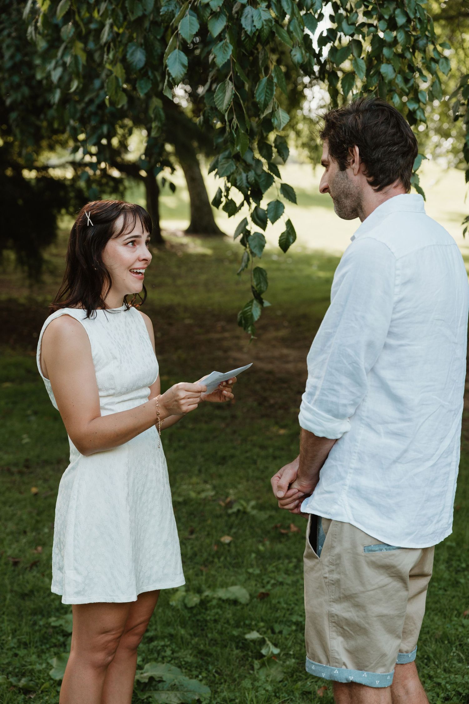 Smiling woman in white dress reads from a card to a man in a sunny green park setting.