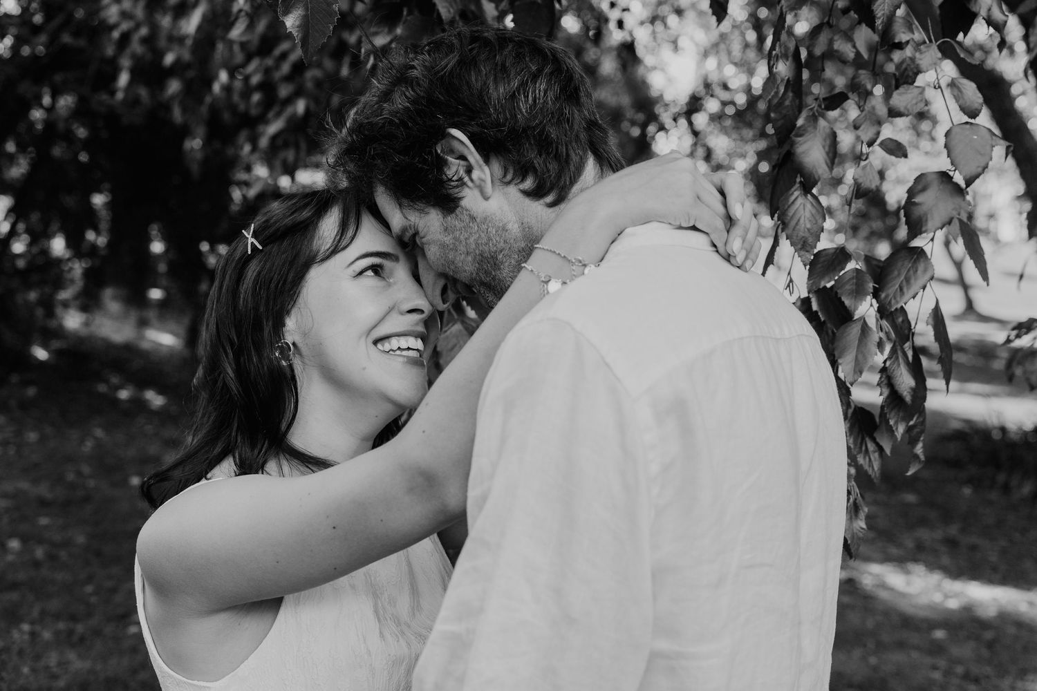 Black and white photo of a smiling couple embracing tenderly under leafy trees in an outdoor setting.