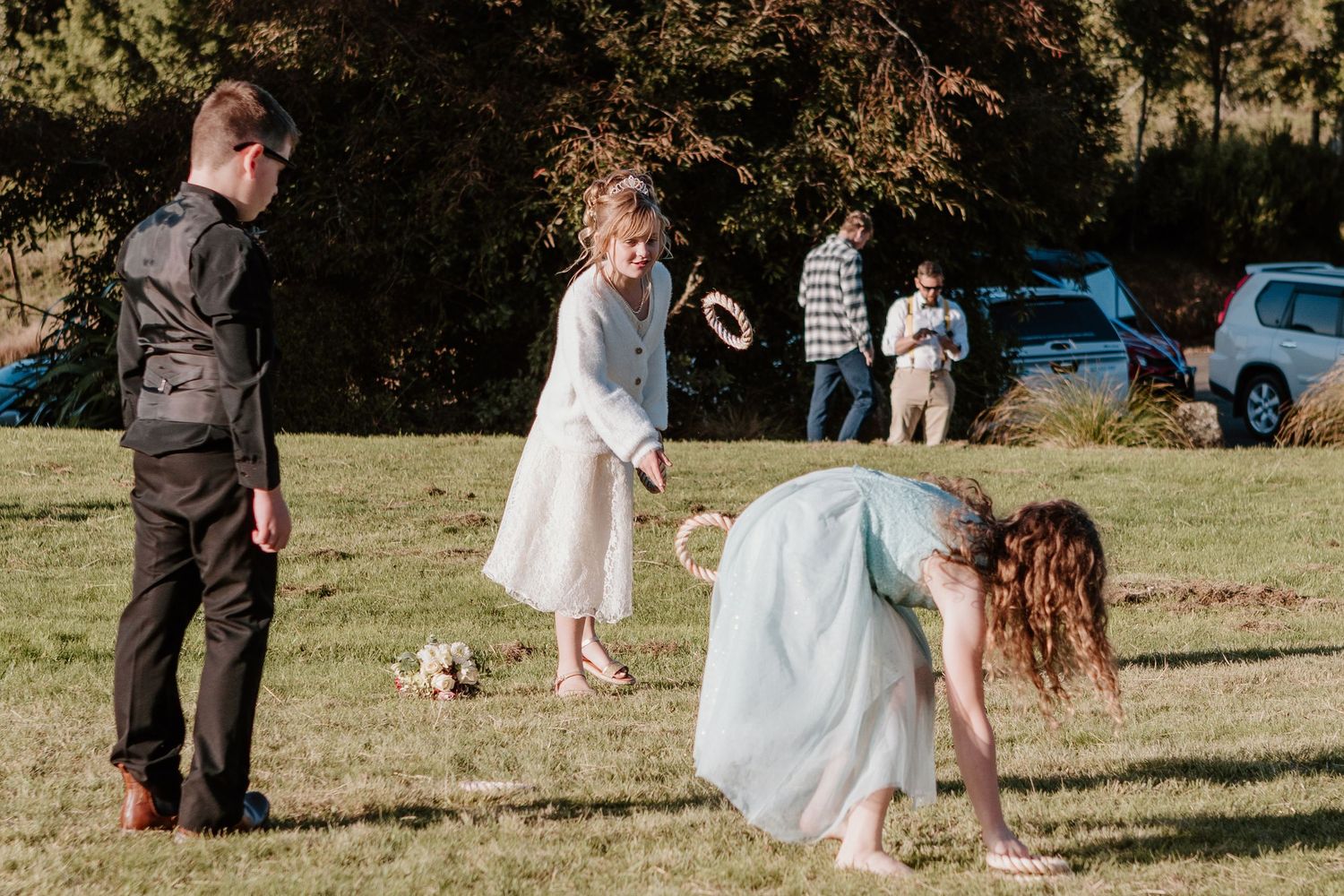 Wedding guests play games and interact on a grassy field with a vintage blue car in the background.